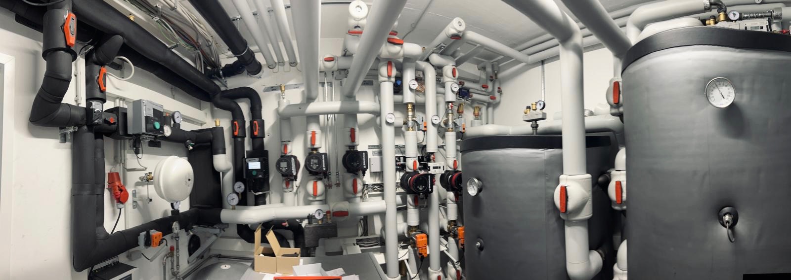 Rozbudowana kotłownia z licznymi rurami, zaworami, pompami obiegowymi i zbiornikami akumulacyjnymi, widoczne także elementy automatyki i instalacji elektrycznej.