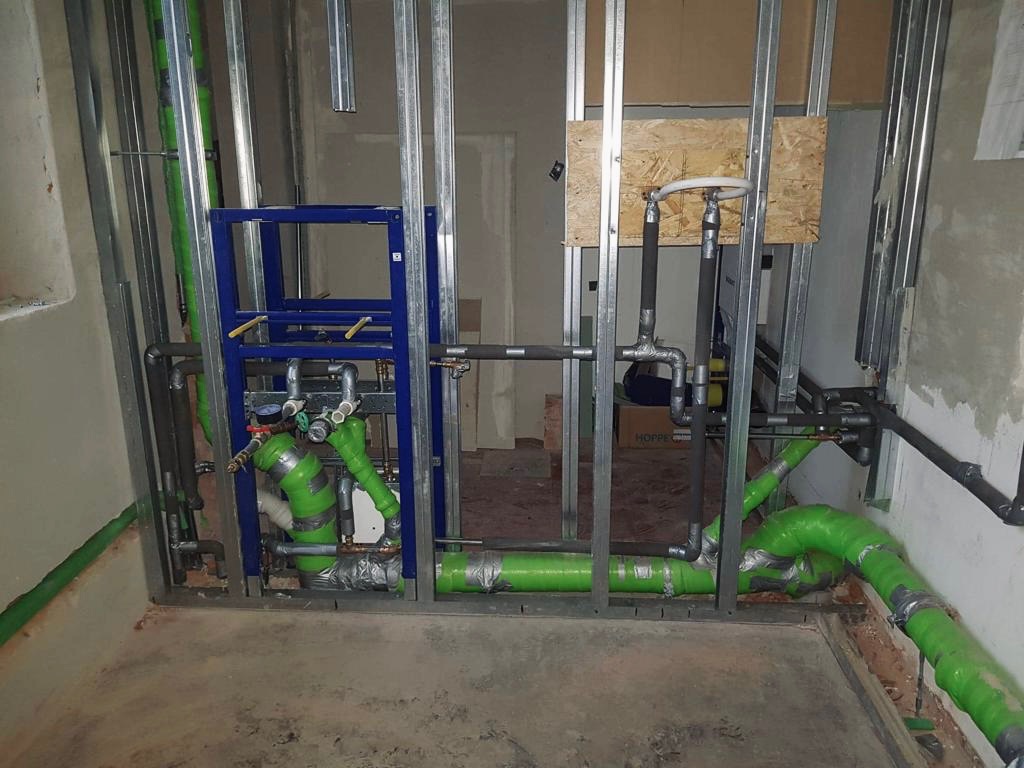 Wnętrze łazienki w trakcie remontu, widoczne stalowe profile ścianek działowych, niebieska rama stelaża podtynkowego WC, rury instalacji wodnej owinięte zieloną folią i szarą taśmą, surowa podłoga...