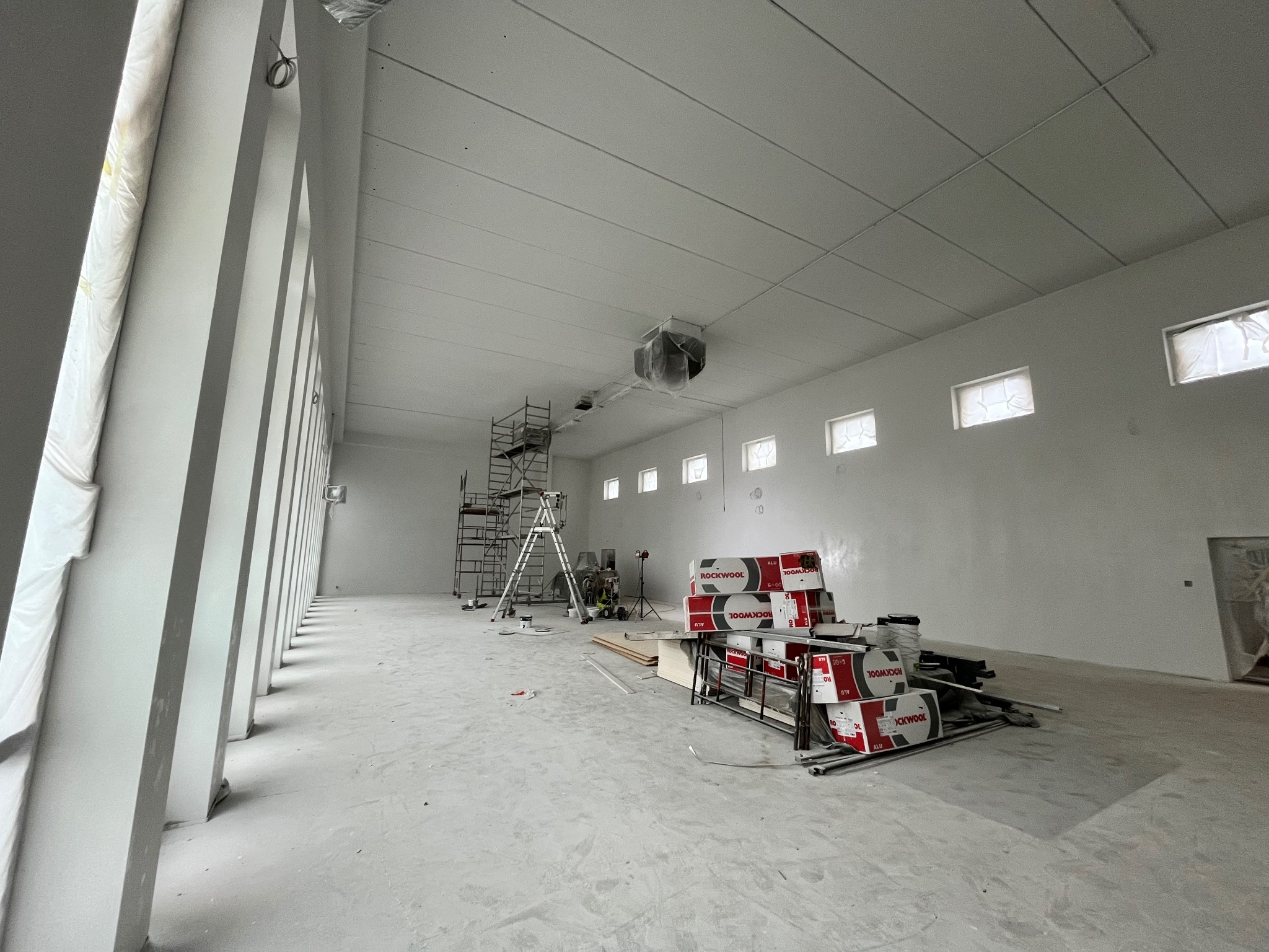 Wnętrze hali w trakcie wykańczania, widoczne rusztowania, puszki z farbą i stos opakowań wełny mineralnej Rockwool. Okna zabezpieczone folią malarską, sufit pomalowany na biało.