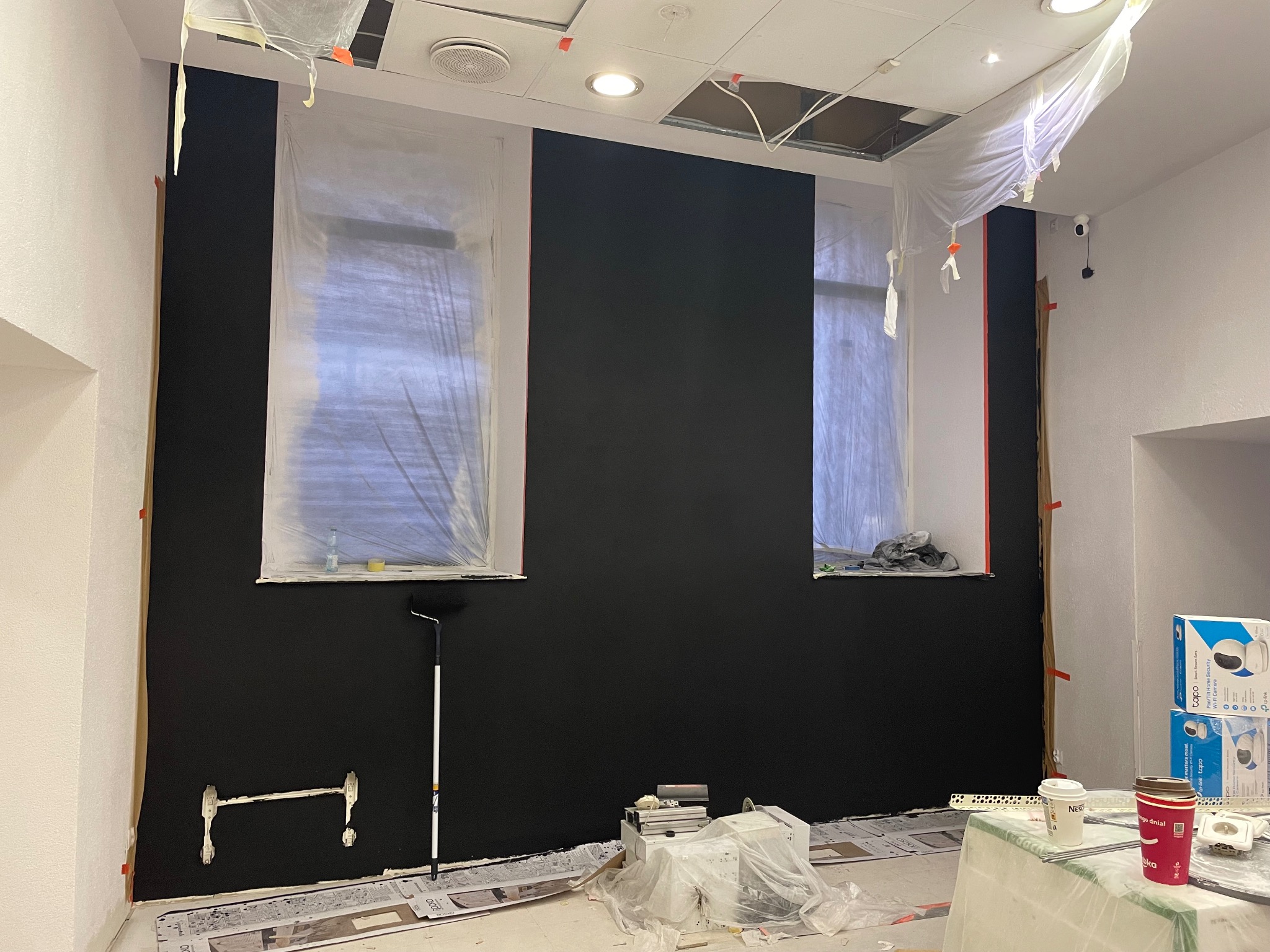 Świeżo pomalowana na czarno ściana w biurze z dwoma oknami zabezpieczonymi folią malarską, widoczne zabezpieczenia folią i taśmą malarską na ścianach, na podłodze rozłożona gazeta malarska, wałek...