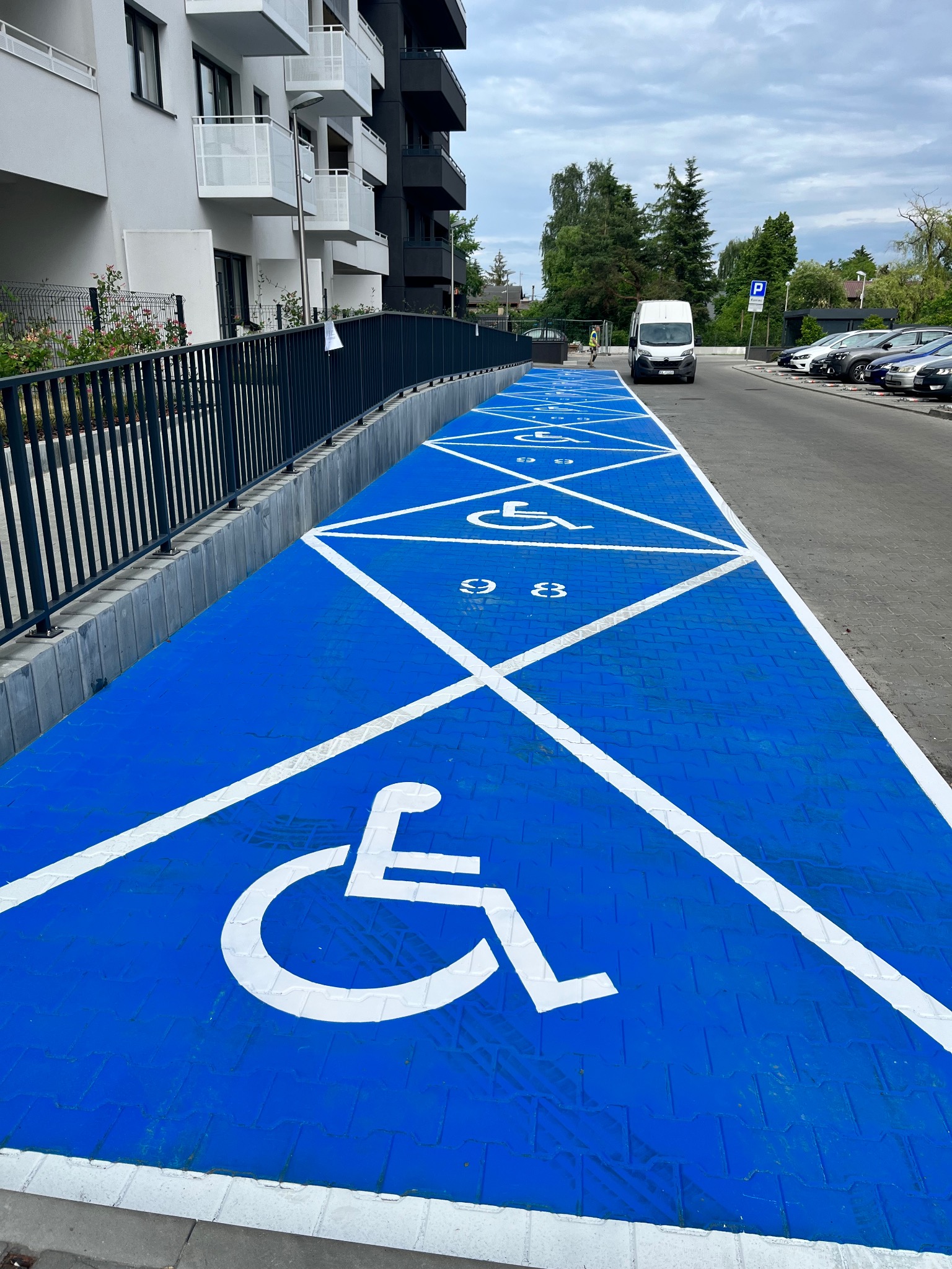 Niebieski parking dla niepełnosprawnych z białymi symbolami i numerami na kostce brukowej, widok z góry w kierunku białego vana i zaparkowanych samochodów.