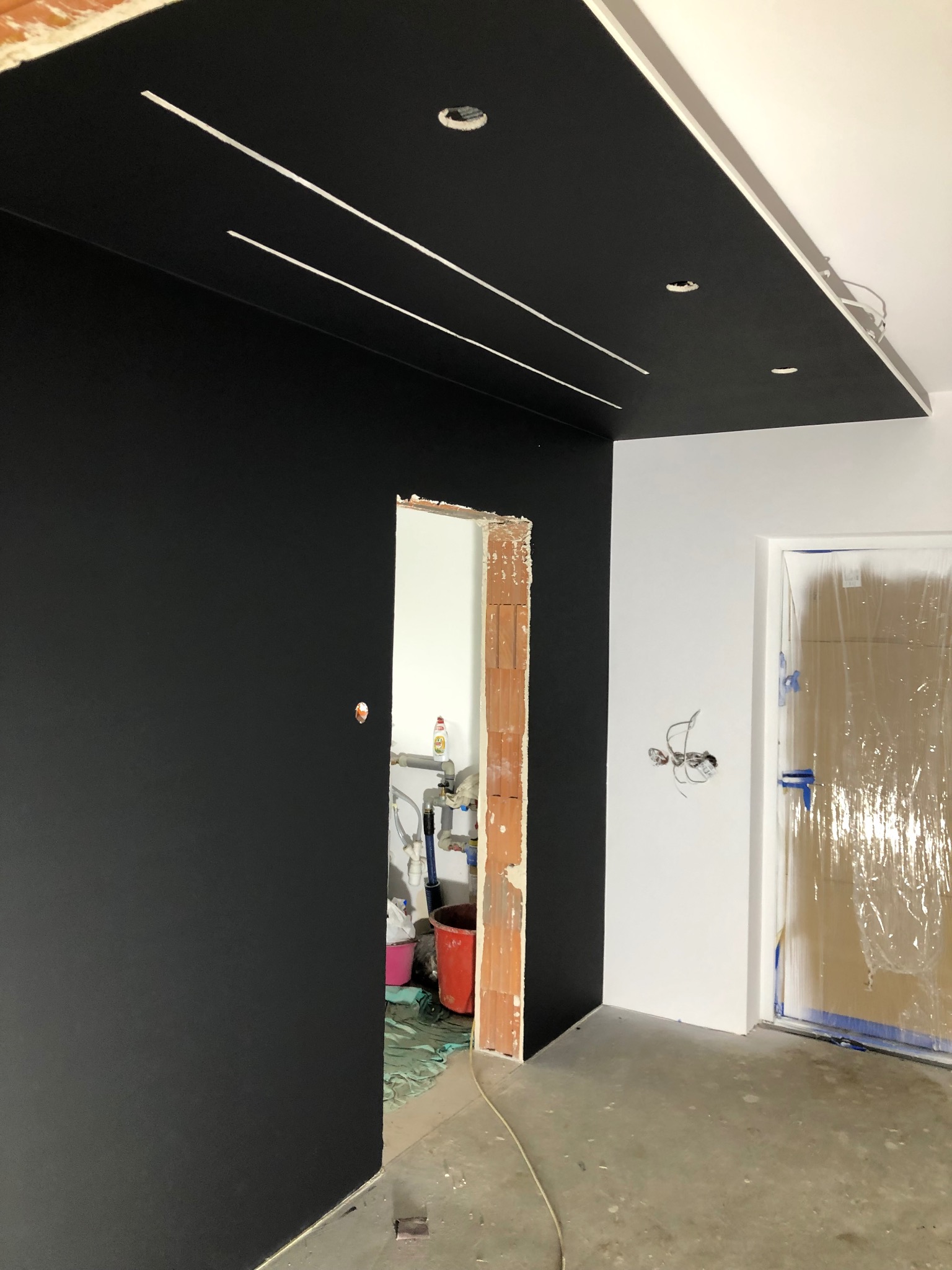 Świeżo pomalowana czarna ściana w pomieszczeniu w trakcie remontu, widoczne przygotowane otwory na oświetlenie w suficie, zabezpieczone folią drzwi oraz fragment ściany z wyprowadzonymi przewodami...