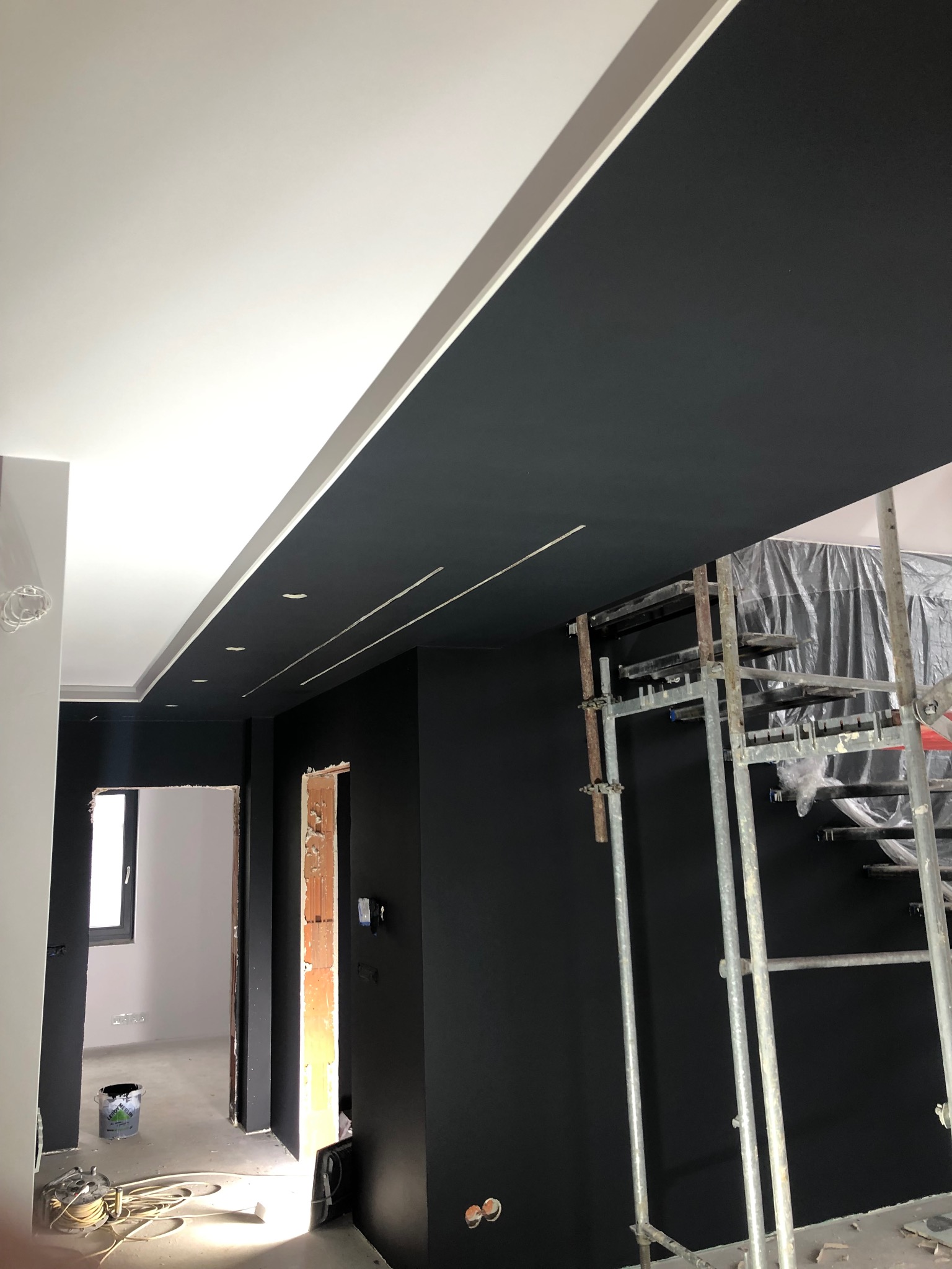 Świeżo pomalowane na czarno ściany i sufit w nowoczesnym wnętrzu, widoczne rusztowanie malarskie z folią ochronną, biały sufit podwieszany z oświetleniem punktowym, przygotowane do dalszych prac...