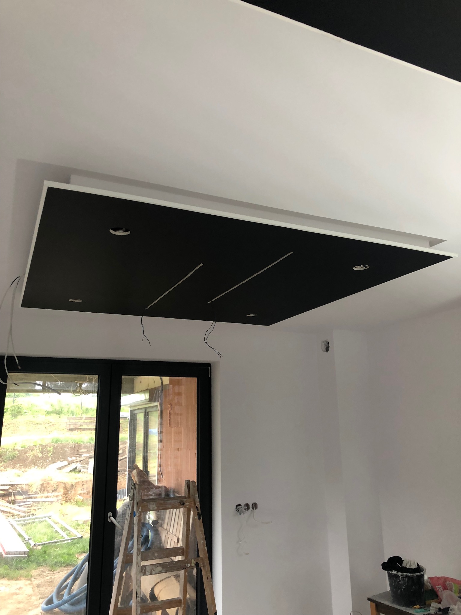 Czarny podwieszany sufit z białym obramowaniem i wpuszczonym oświetleniem w trakcie instalacji, widoczne wystające kable, drabina oparta o okno z widokiem na plac budowy.