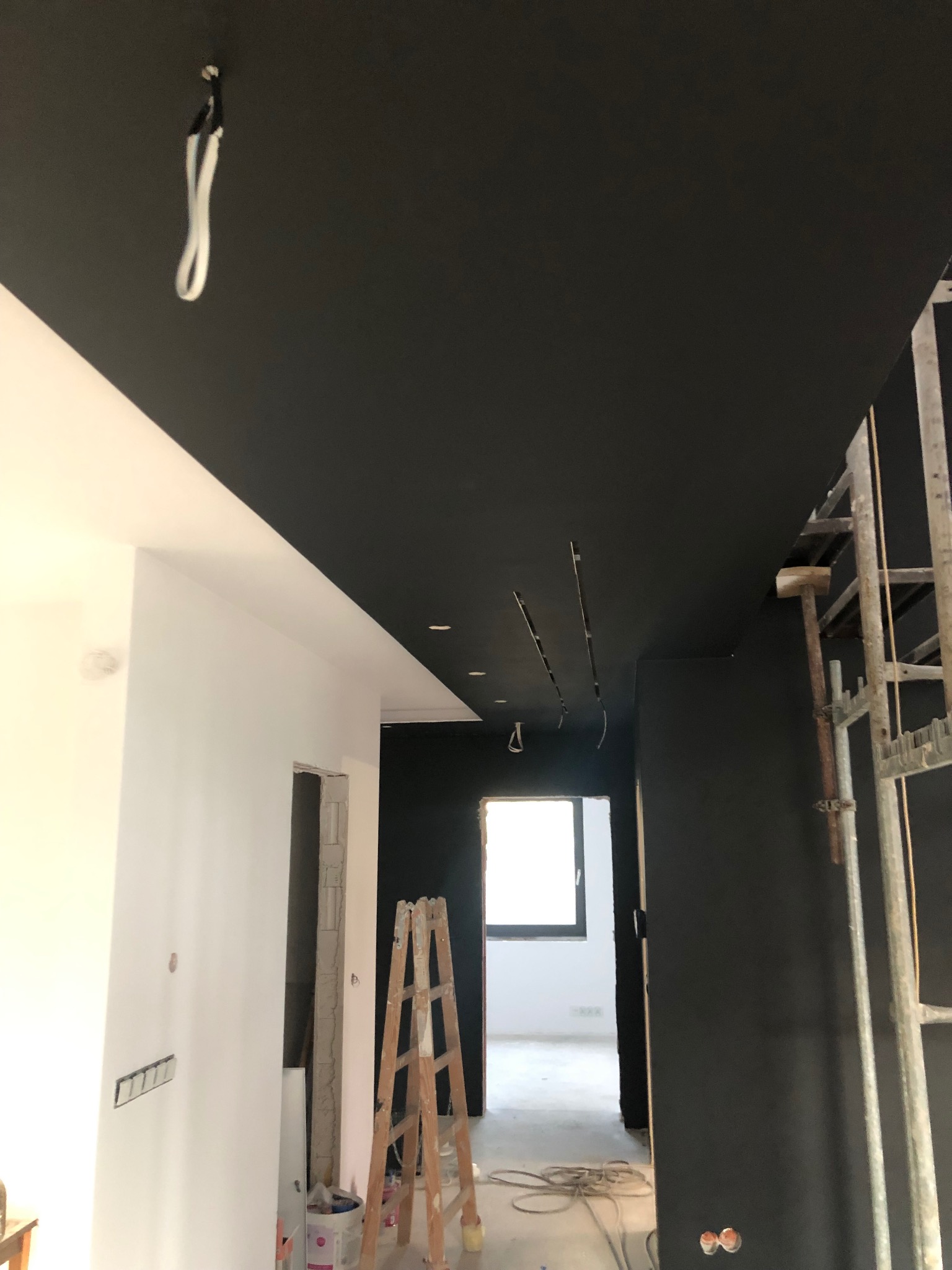 Wnętrze w trakcie remontu: czarny sufit z wystającymi kablami, białe ściany, drabina oparta o ścianę, widoczne ślady po gniazdkach elektrycznych, metalowe rusztowanie przy ścianie, okno w tle.