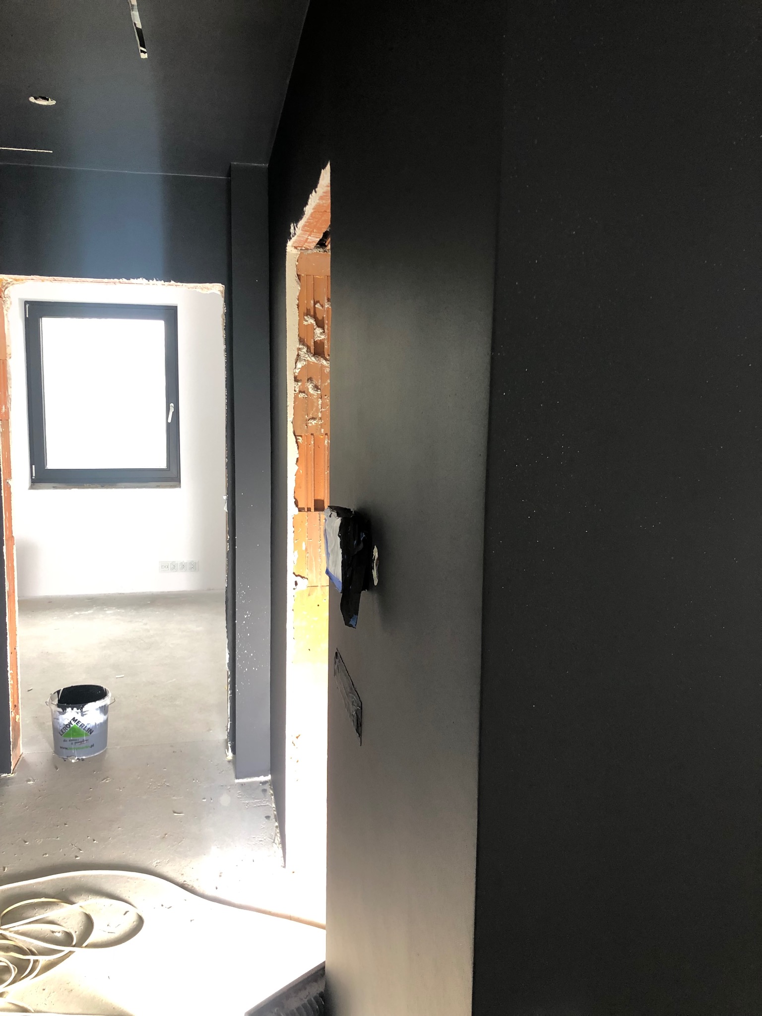 Wnętrze w trakcie malowania na ciemny kolor w Michałowicach, widoczna ściana po malowaniu, wiadro z farbą i fragment odsłoniętej cegły przy framudze drzwi.