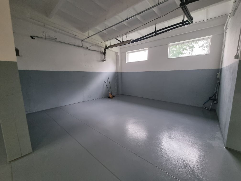 Pomalowane na szaro pomieszczenie gospodarcze z dwoma oknami, widoczne rury pod sufitem i grzejnik przy ścianie. Na podłodze widoczne ślady po malowaniu. W rogu stoi miotła.