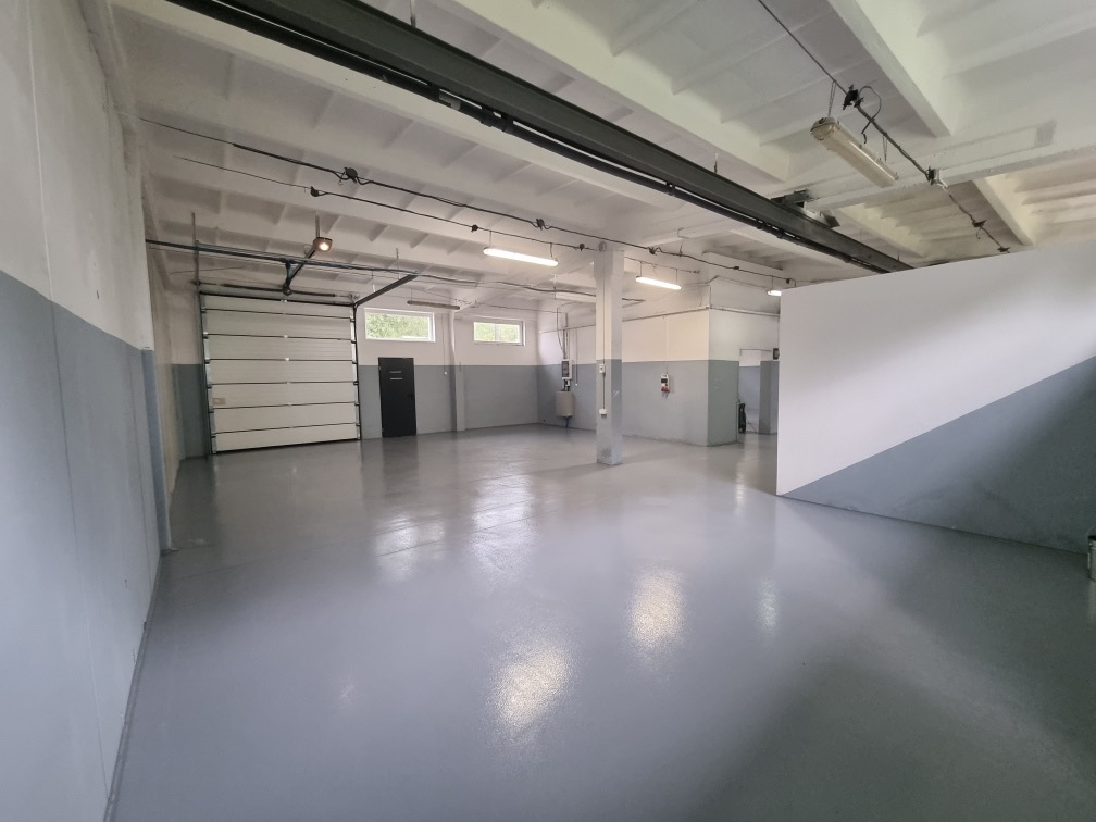 Wnętrze hali przemysłowej po malowaniu, z szarą podłogą z żywicy epoksydowej, biało-szare ściany i biały sufit z widocznymi instalacjami, brama garażowa segmentowa w tle.