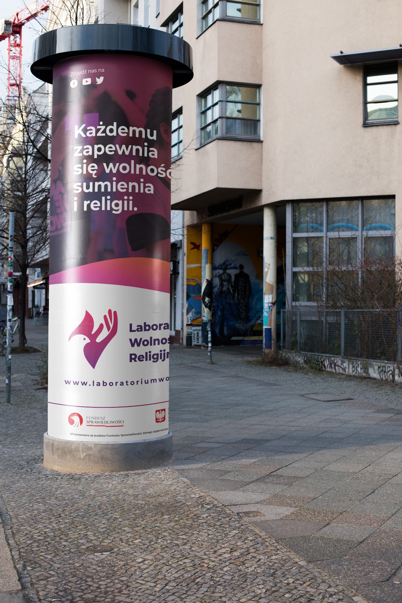 Słup ogłoszeniowy z komunikatem 'Każdemu zapewnia się wolność sumienia i religii' oraz logo Laboratorium Wolności, umieszczony na chodniku w przestrzeni miejskiej.