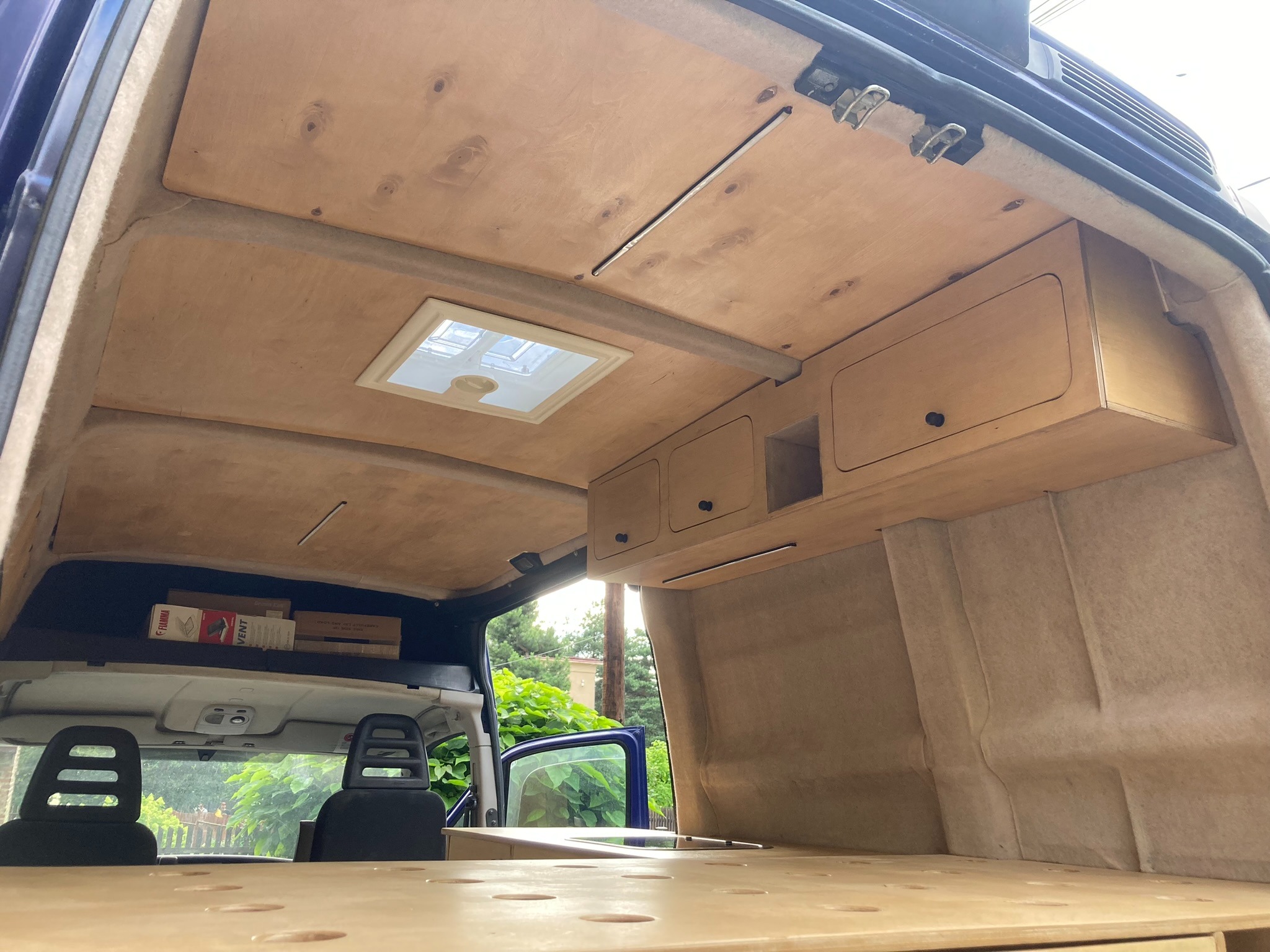 Wnętrze kampera w trakcie zabudowy: sklejka na ścianach i suficie, zabudowa szafek z drewna, okno dachowe, widoczne pudełka na półce nad siedzeniami.