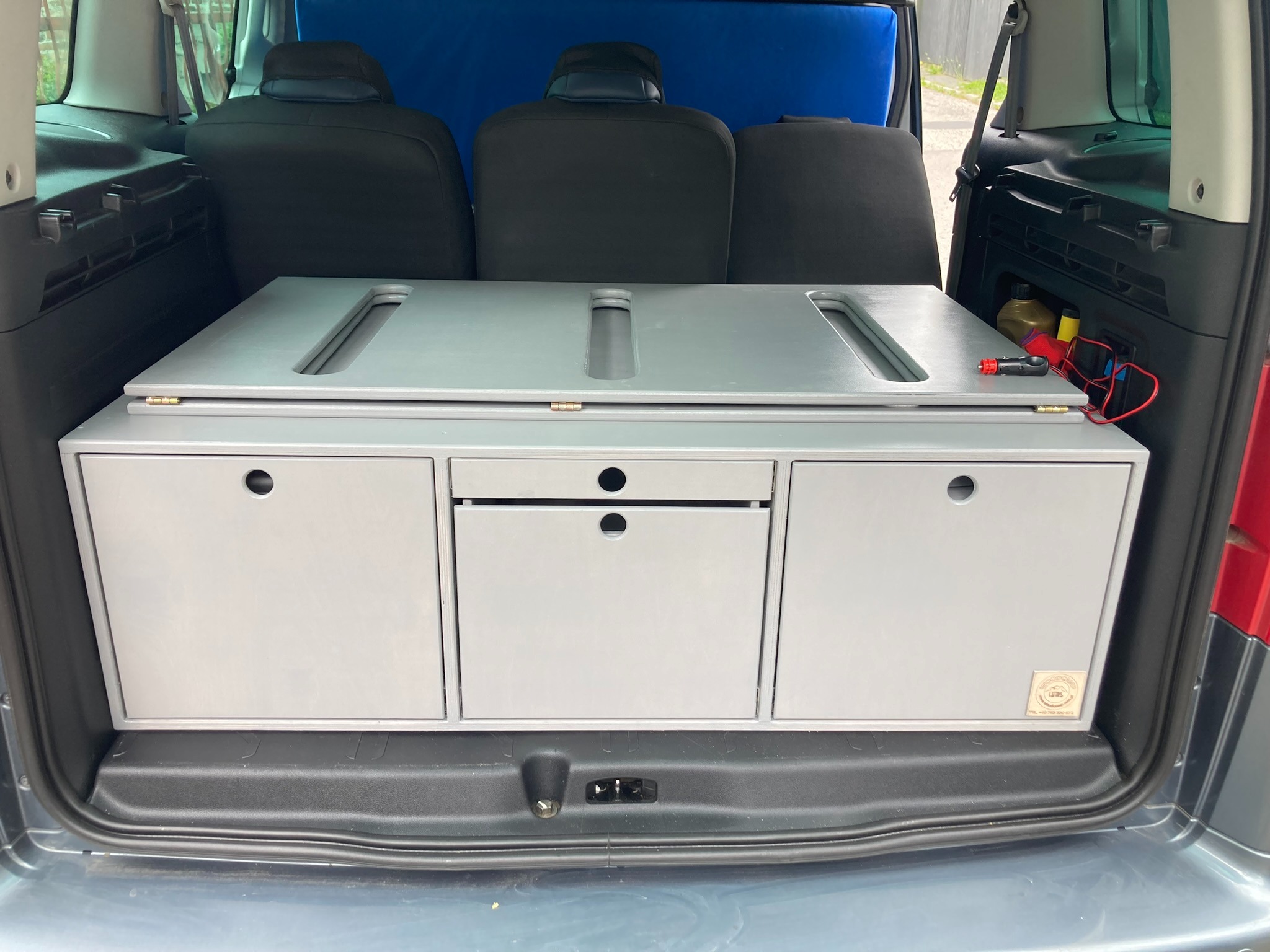 Zabudowa modułowa z szufladami i blatem w bagażniku samochodu typu van, z widocznym okablowaniem i akcesoriami po prawej stronie.