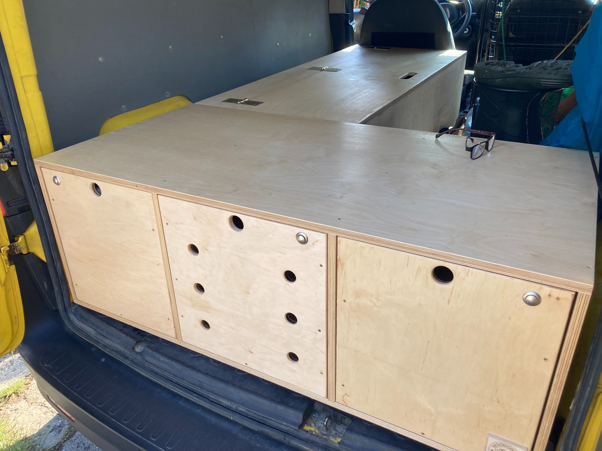 Wnętrze żółtego vana z zabudową z jasnej sklejki: szafki z okrągłymi otworami i blat, na którym leżą okulary.