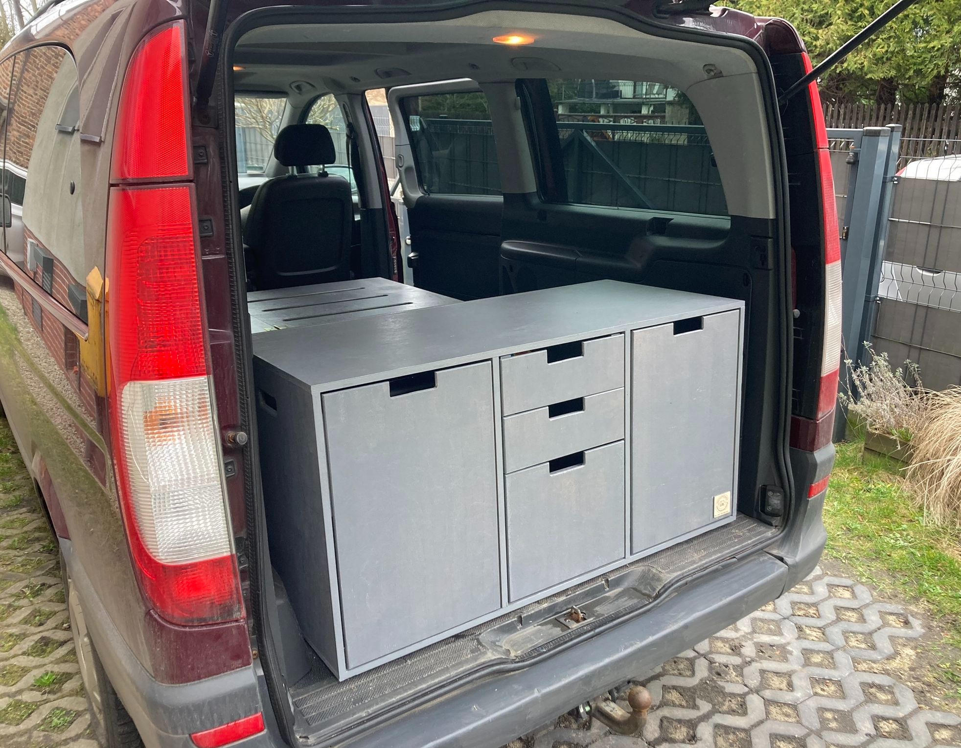 Szare modułowe meble z szufladami zamontowane w bagażniku samochodu typu van, widoczne z zewnątrz na tle kostki brukowej i ogrodzenia.