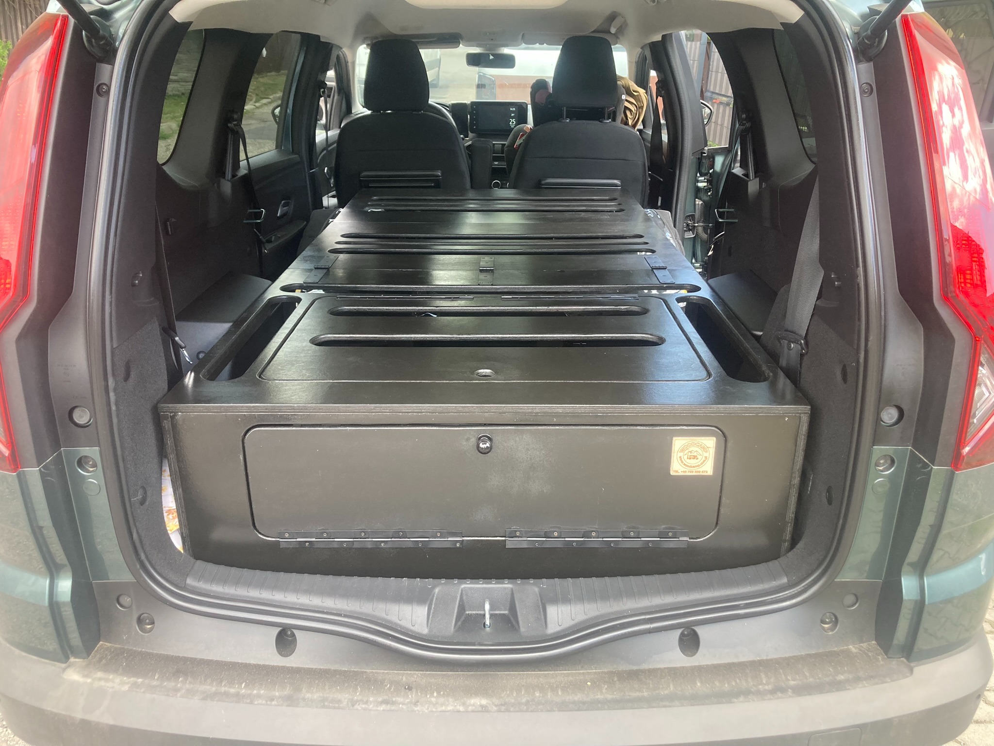 Czarna zabudowa modułowa z szufladami w bagażniku samochodu typu kombi, widoczna od strony otwartej klapy bagażnika, z naklejką producenta na froncie.