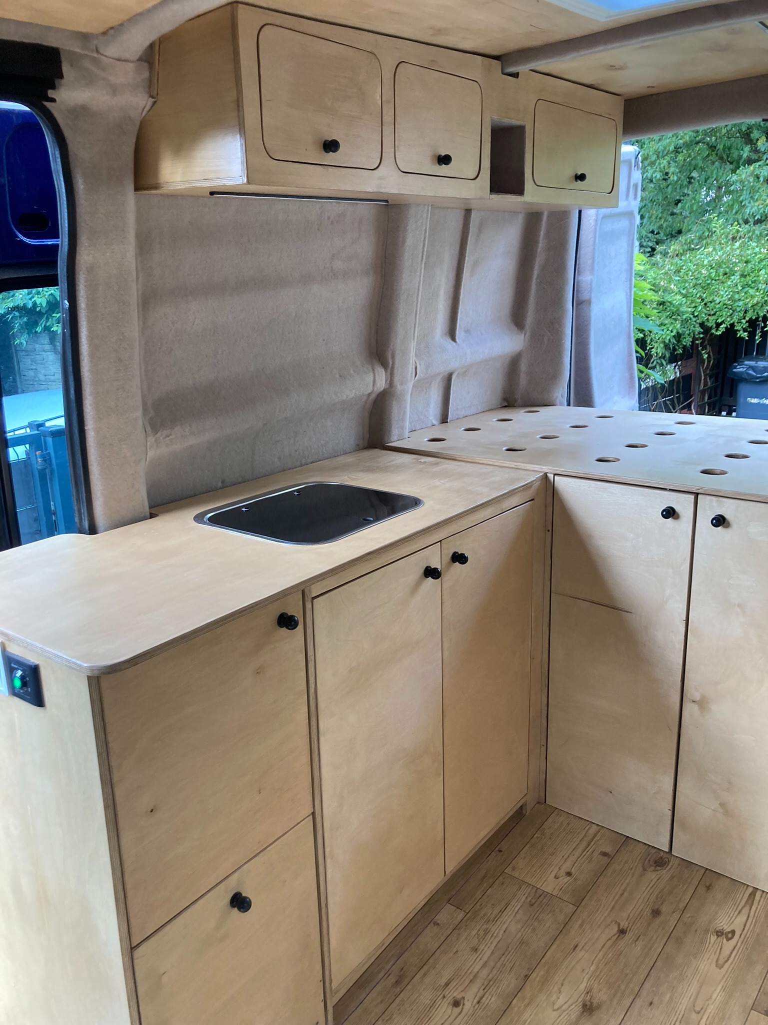 Wnętrze kampera z zabudową kuchenną z jasnego drewna, obejmującą szafki wiszące i dolne, zlew w blacie oraz blat z otworami, widoczne okno i elementy karoserii pojazdu.