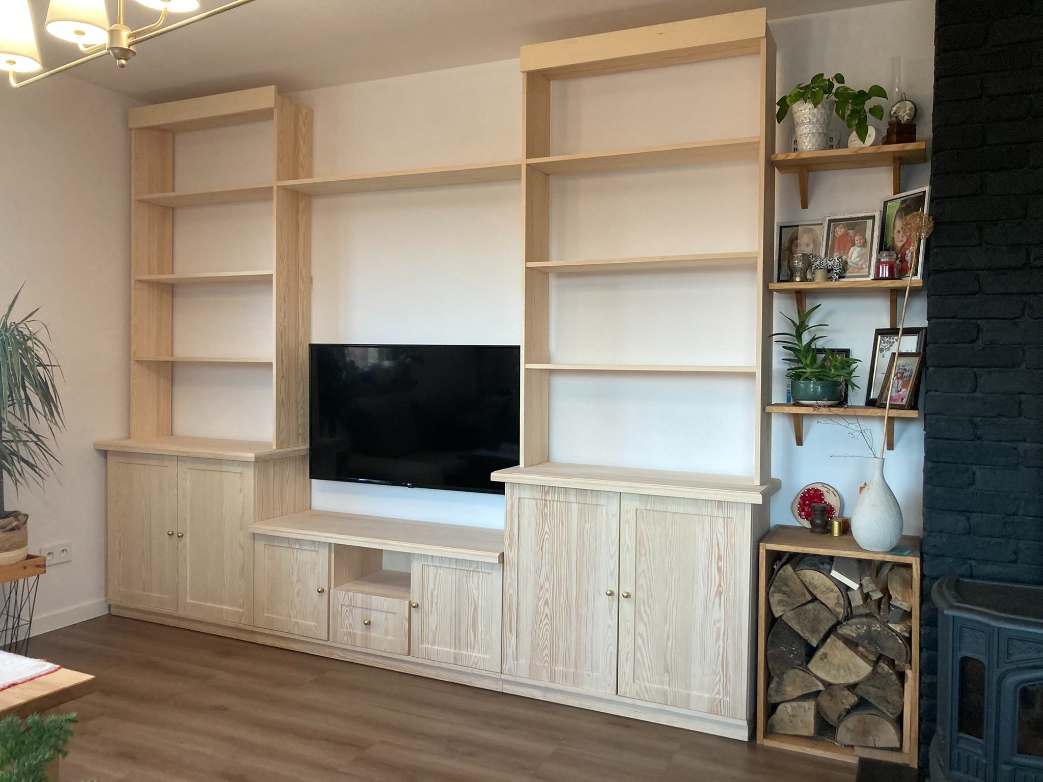 Jasna, drewniana zabudowa ściany z półkami i szafkami, z miejscem na telewizor, uzupełniona dekoracjami na półkach i obok kominka.