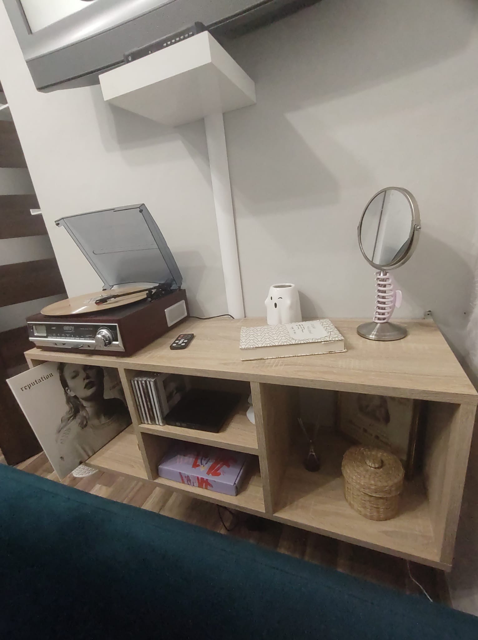 Jasna, drewnopodobna komoda z półkami, na której stoi gramofon z płytą, książka, lusterko na sprężynie i dekoracje.