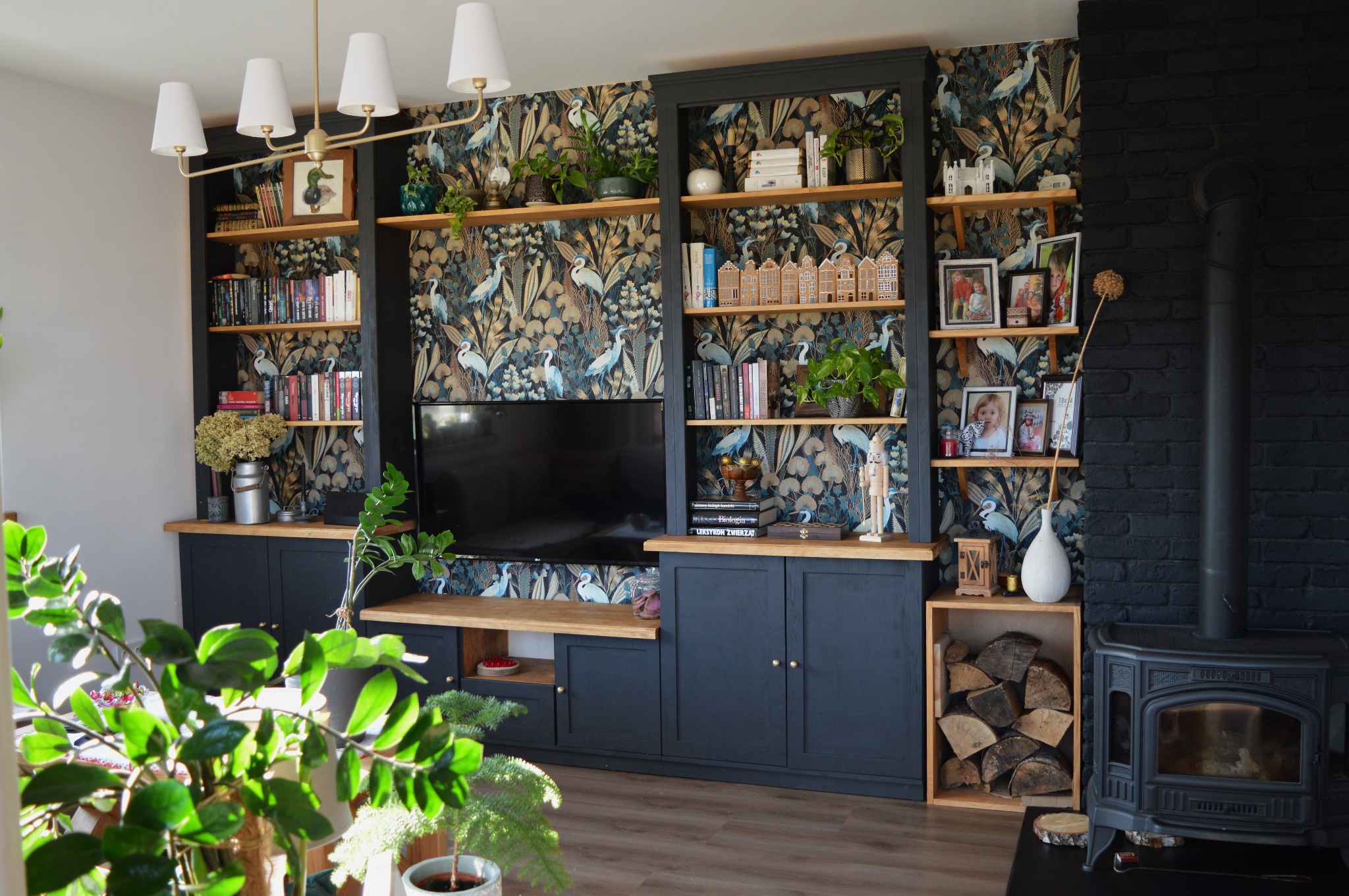 Ciemna szafa na wymiar z drewnianymi półkami, ozdobiona książkami, roślinami, zdjęciami i dekoracjami, na tle tapety w ptaki i ceglanej ściany z piecem.