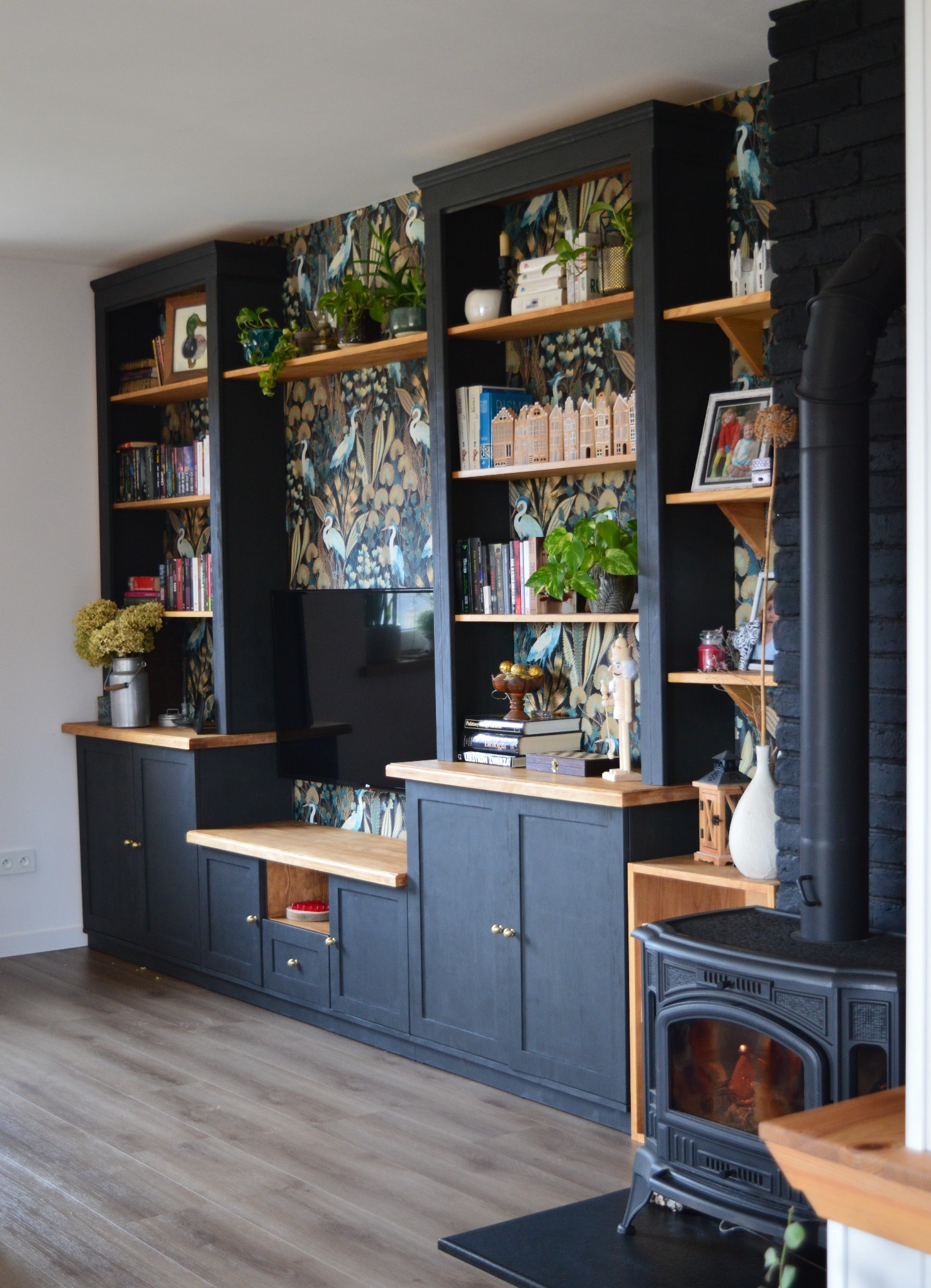Ciemna zabudowa meblowa z drewnianymi blatami i otwartymi półkami, na których stoją książki, rośliny i dekoracje, z telewizorem pośrodku, na ścianie wzorzysta tapeta, obok piec wolnostojący.