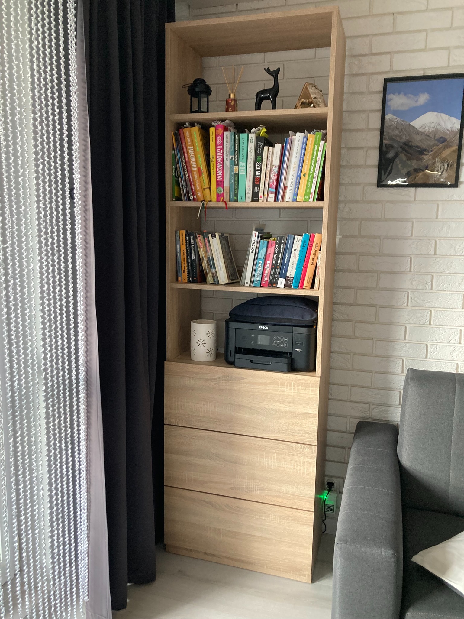 Regał z jasnego drewna z trzema szufladami i półkami wypełnionymi książkami, dekoracjami i drukarką, ustawiony przy ceglanej ścianie obok zasłon w kolorze czarnym i białym.