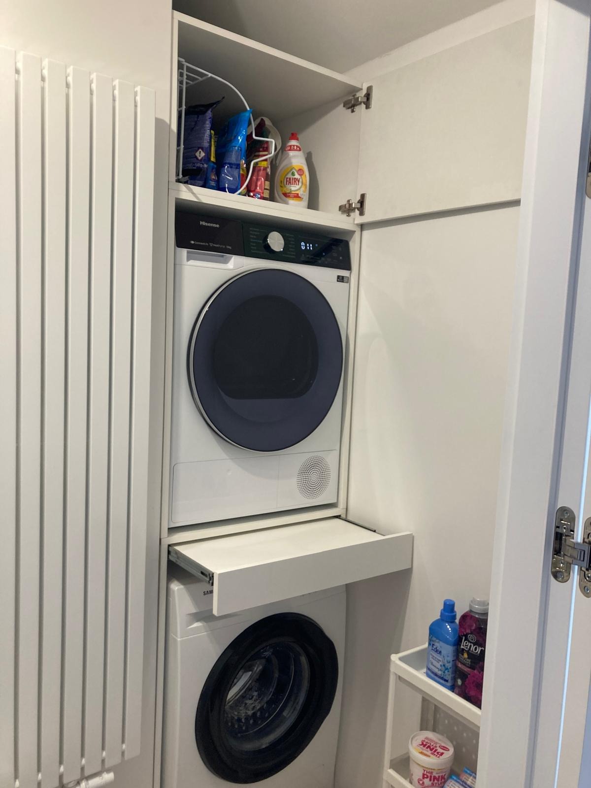 Biała pralka i suszarka w zabudowie meblowej z wysuwanym blatem, obok grzejnik panelowy i półki z detergentami.