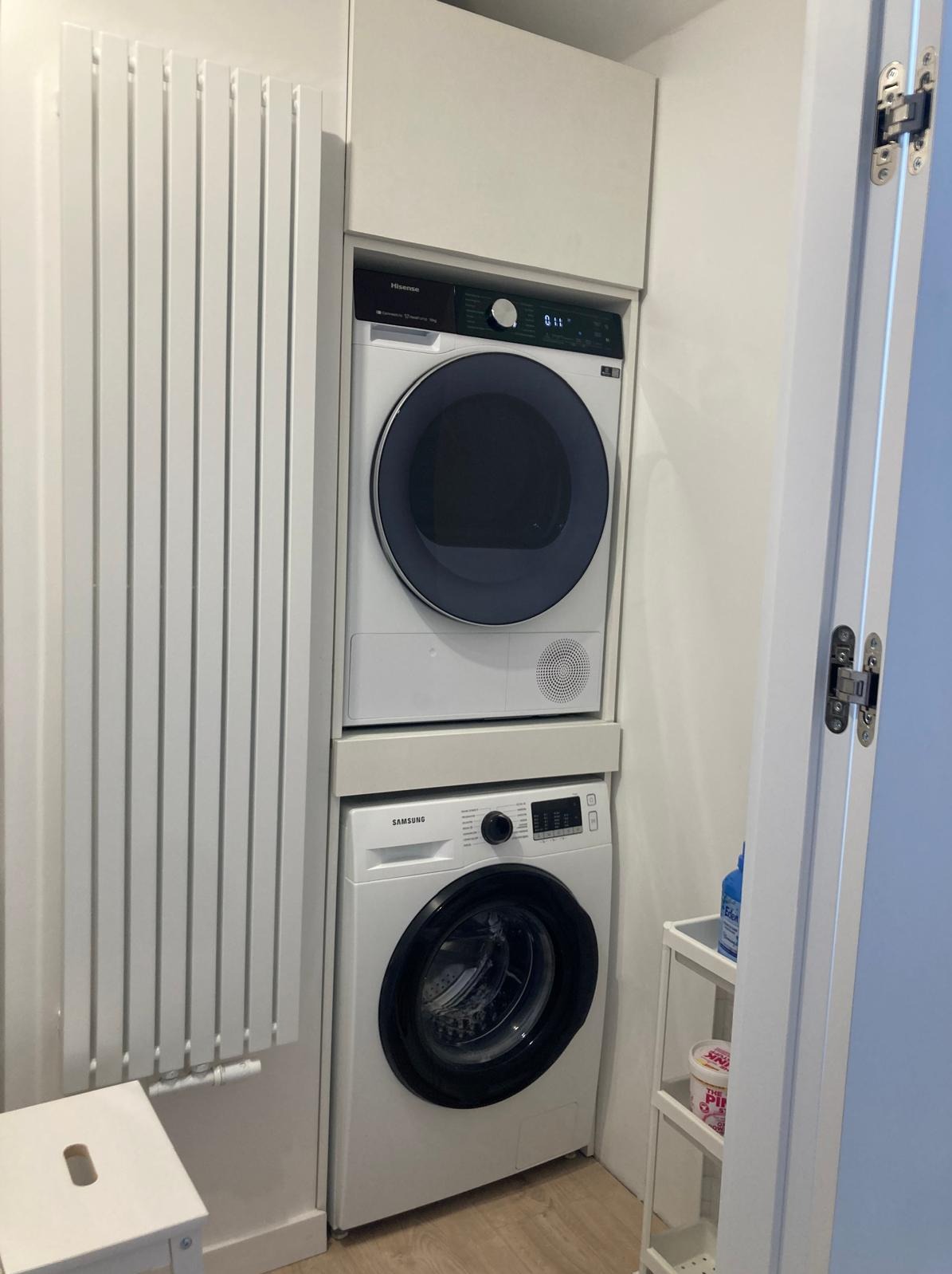 Pralka i suszarka w zabudowie w Katowicach, obok grzejnik panelowy i mały regał z detergentami.