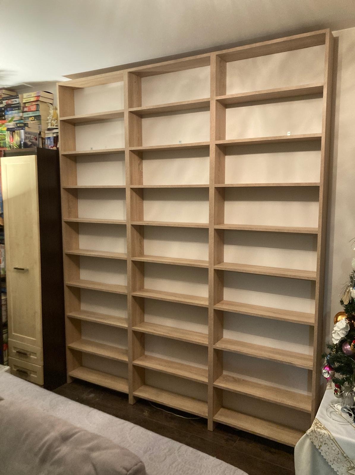 Trzyczęściowa, wysoka, drewniana konstrukcja regału z pustymi półkami, przylegająca do ściany w pomieszczeniu z elementami dekoracji świątecznej w tle.