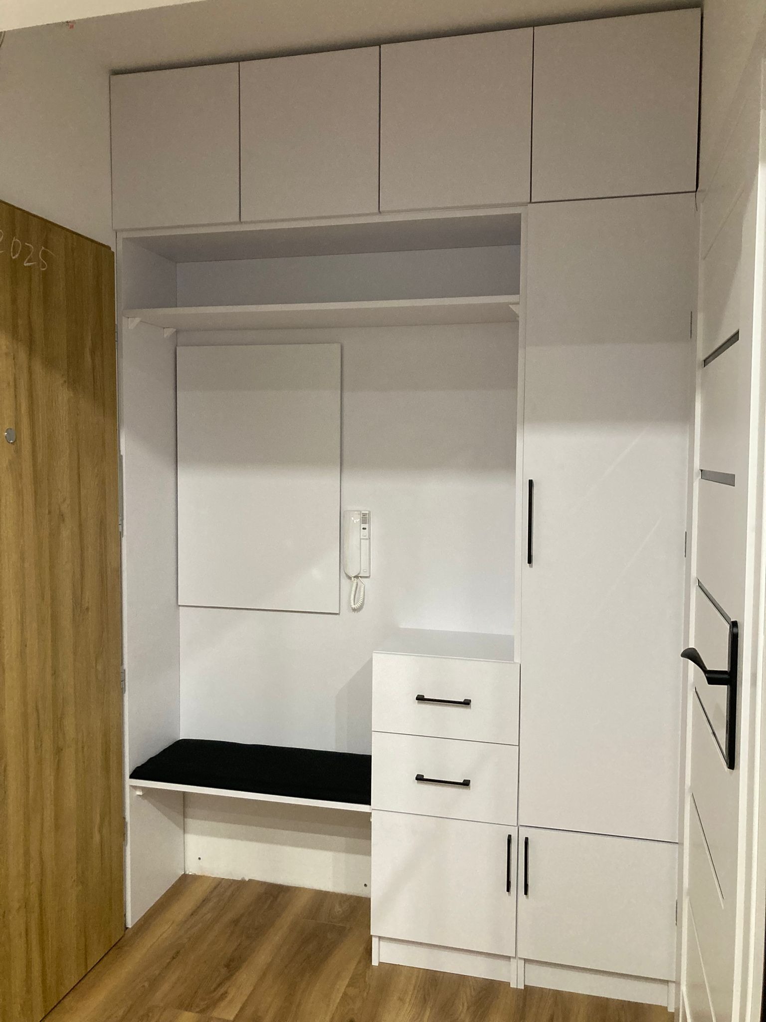 Biała zabudowa meblowa w przedpokoju z siedziskiem, szafkami górnymi i dolnymi oraz lustrem, dopasowana do wnęki.