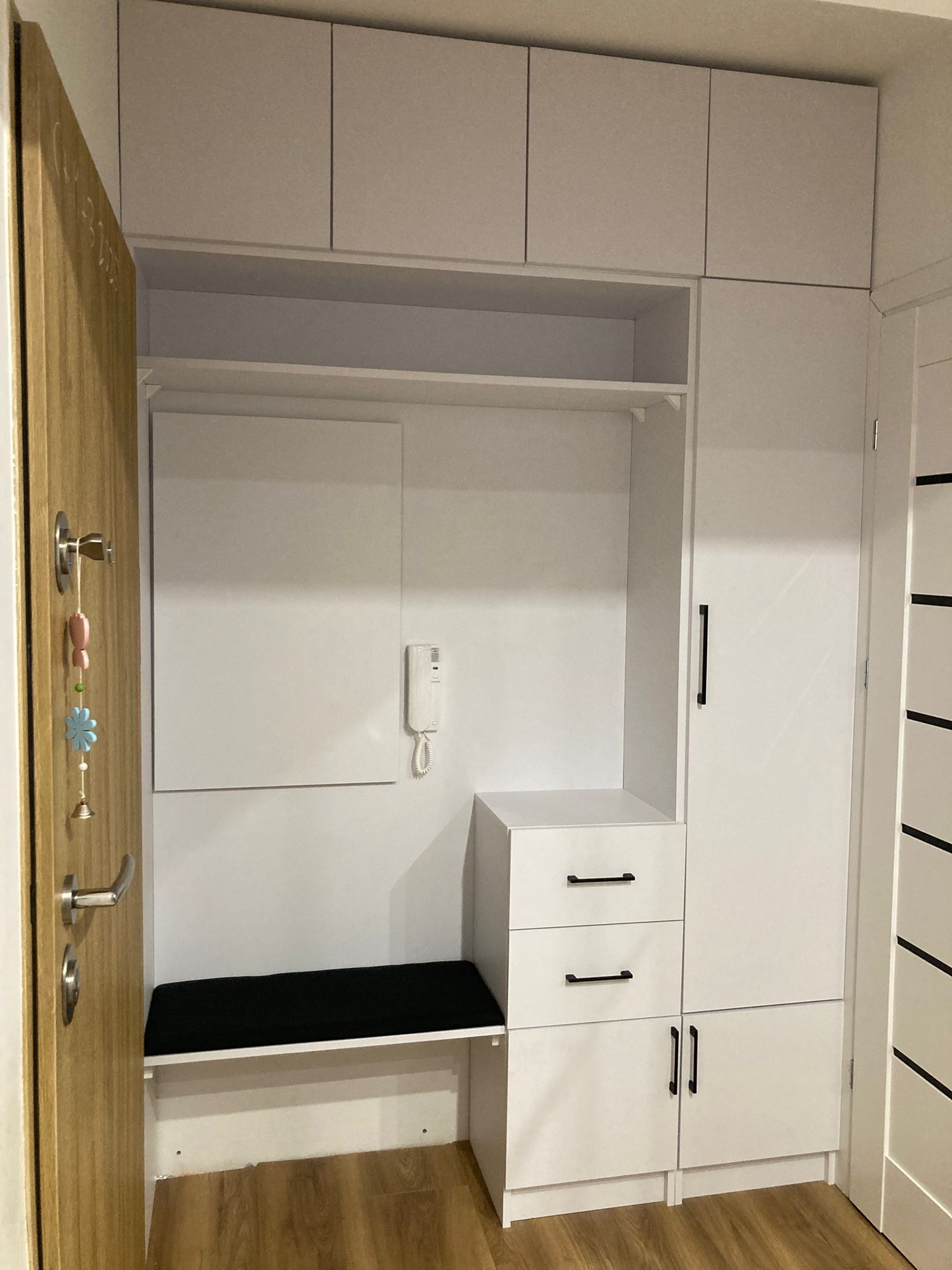 Biała zabudowa meblowa w przedpokoju z siedziskiem, szafkami górnymi i dolnymi oraz wieszakiem, uzupełniona o domofon na ścianie i drewniane drzwi wejściowe z ozdobnym wisiorkiem.