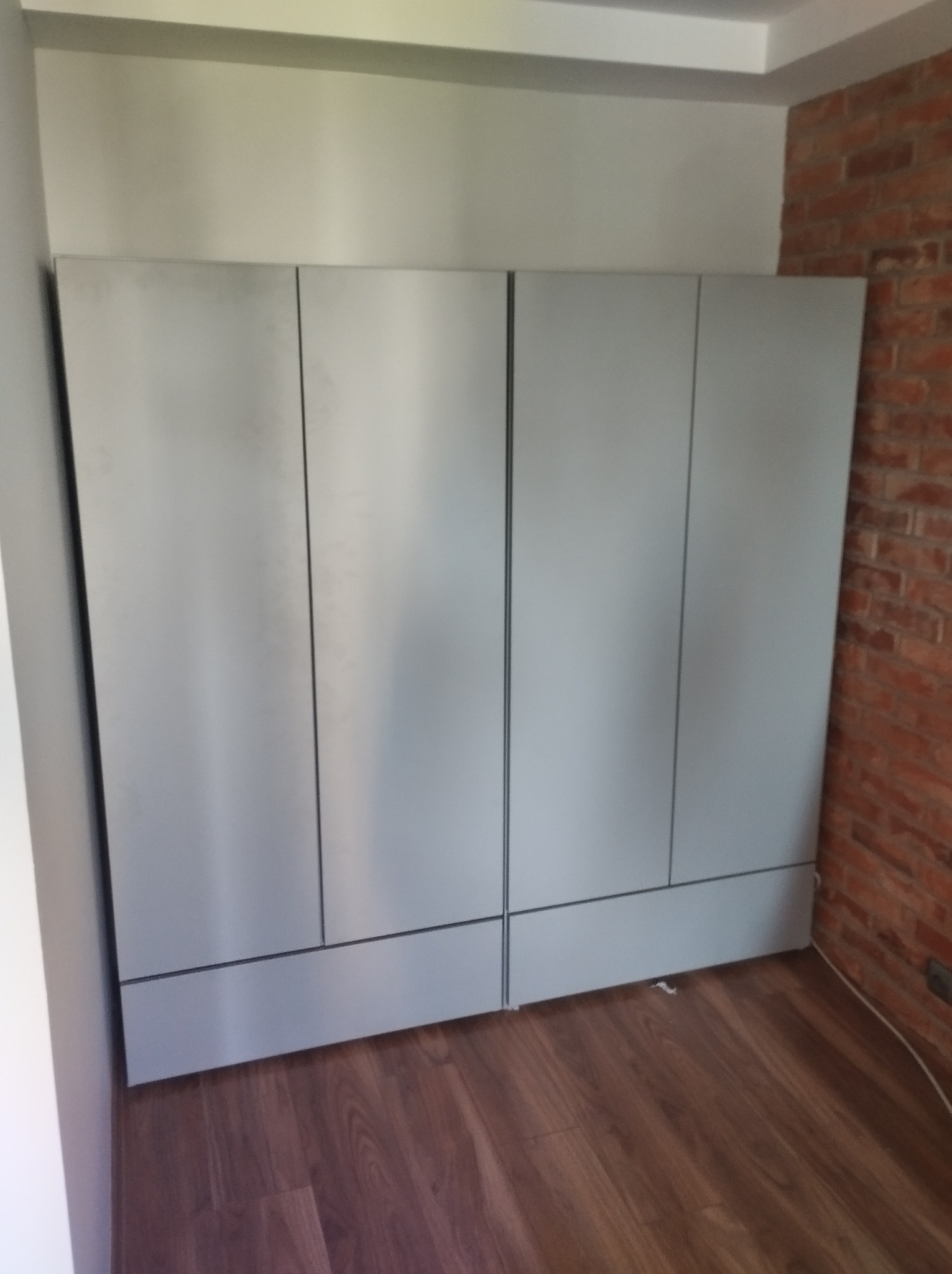 Czterodrzwiowa szafa wnękowa z gładkimi, szarymi frontami, umieszczona w rogu pokoju z fragmentem ściany wykończonej czerwoną cegłą.