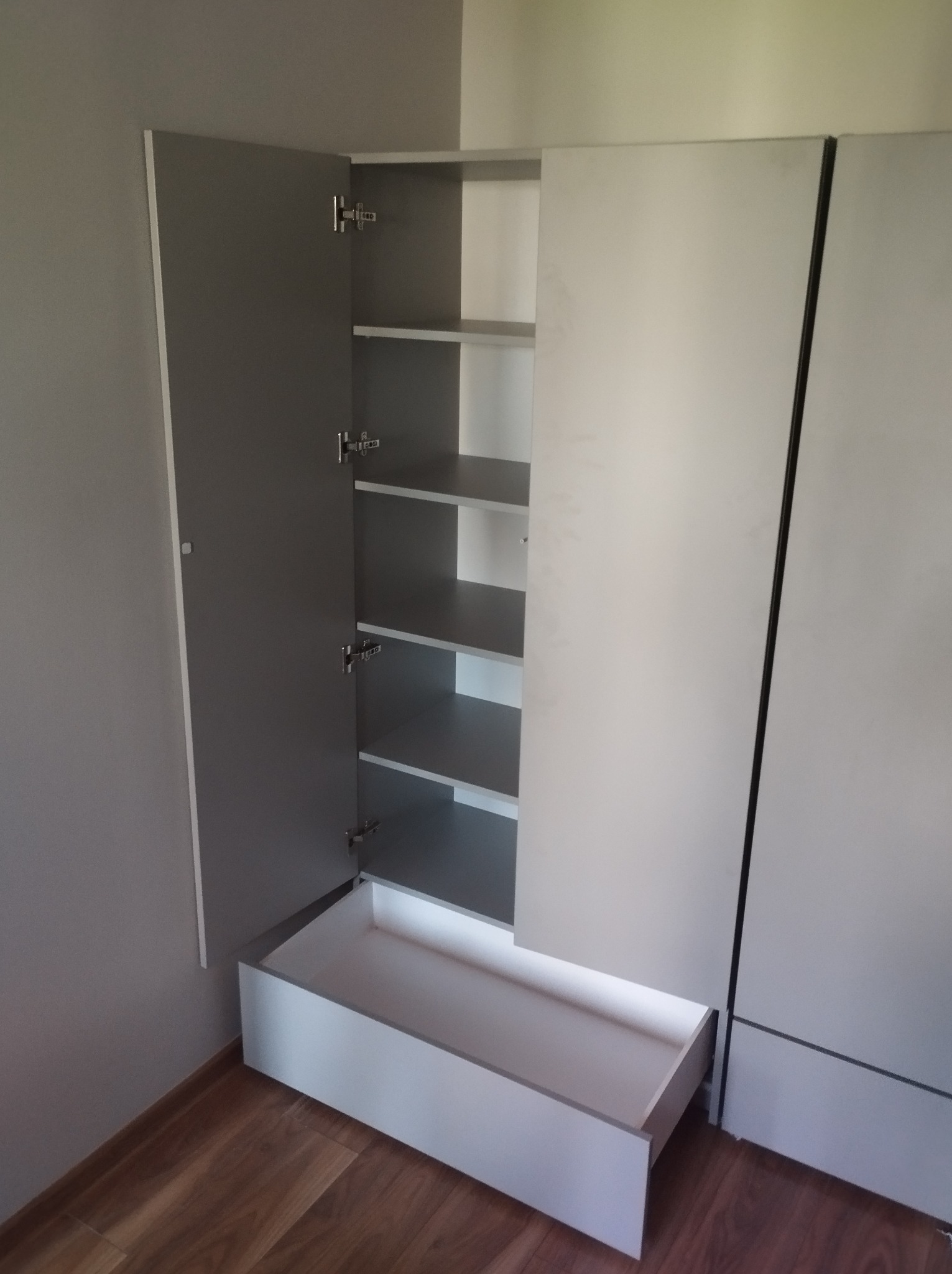 Szara szafa z otwartymi drzwiami, ukazująca półki i wysuniętą szufladę, na tle drewnianej podłogi i szarej ściany.