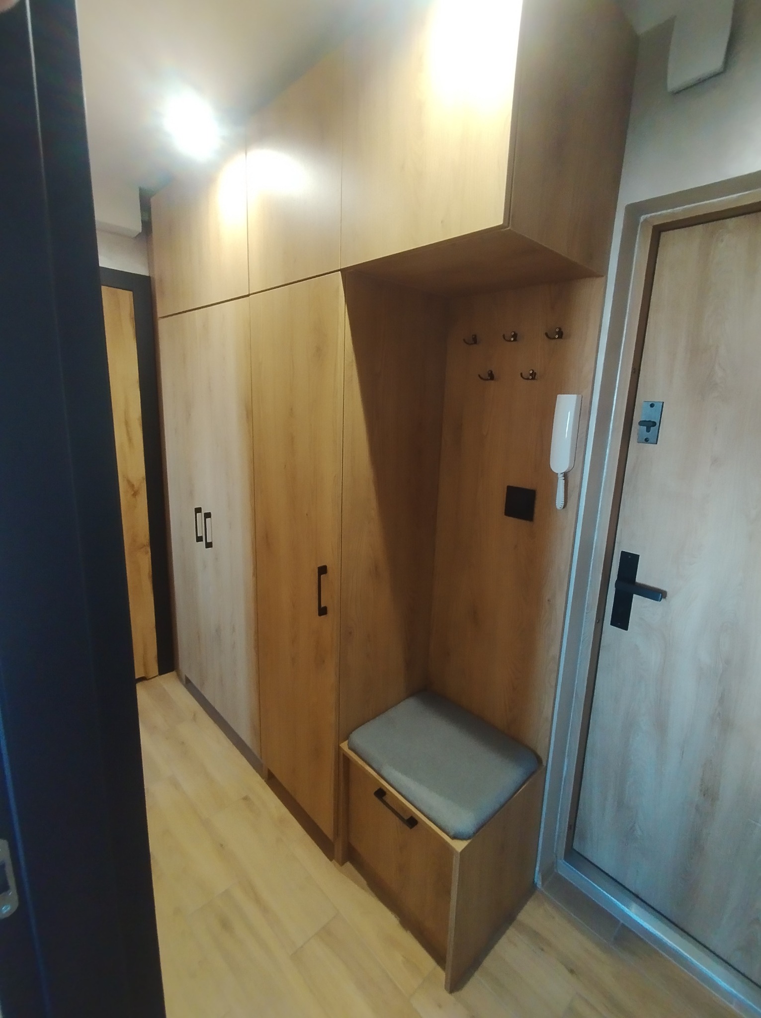 Zabudowa przedpokoju z szafami z drewnopodobnej płyty, siedziskiem ze schowkiem i wieszakami na ścianie, obok drzwi wejściowe.