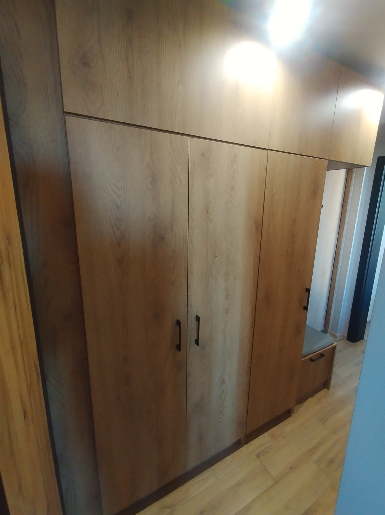 Szafa wnękowa z drewnianymi frontami i czarnymi uchwytami, zintegrowana z siedziskiem w przedpokoju, widok z perspektywy podłogi.