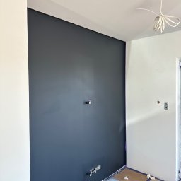 SZYM-PRO CONSTRUCTION - Świeżo pomalowana ściana na ciemny grafit w narożniku pokoju, z widocznymi puszkami elektrycznymi i zabezpieczoną podłogą, przygotowana do dalszych prac wykończeniowych.