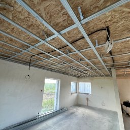 SZYM-PRO CONSTRUCTION - Wnętrze w trakcie montażu metalowej konstrukcji sufitu podwieszanego na płycie OSB, widoczne kable elektryczne i przygotowane ściany do dalszych prac.