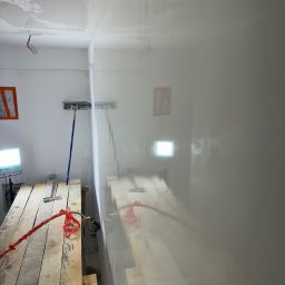 SZYM-PRO CONSTRUCTION - Wnętrze w trakcie prac wykończeniowych: świeżo nałożona, mokra warstwa gładzi na ścianach i suficie, widoczne rusztowanie robocze z narzędziami, lampa oświetleniowa i szpachla na kiju.