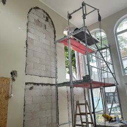 SZYM-PRO CONSTRUCTION - Wnętrze w trakcie prac murarskich: w ścianie wycięte otwory w kształcie łuków, widoczne bloki z betonu komórkowego, rusztowanie, drabina i narzędzia.