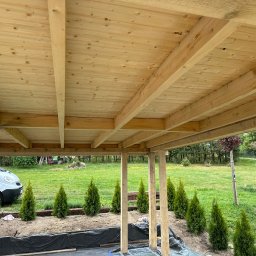 SZYM-PRO CONSTRUCTION - Surowy drewniany strop z widocznymi belkami, częściowo zadaszony, widok od dołu, z zewnątrz widoczne tuje i biały samochód.