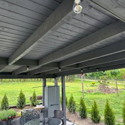 SZYM-PRO CONSTRUCTION - Szare drewniane zadaszenie tarasu z widokiem na ogród, oświetlone żarówkami, z elementami wyposażenia na pierwszym planie.