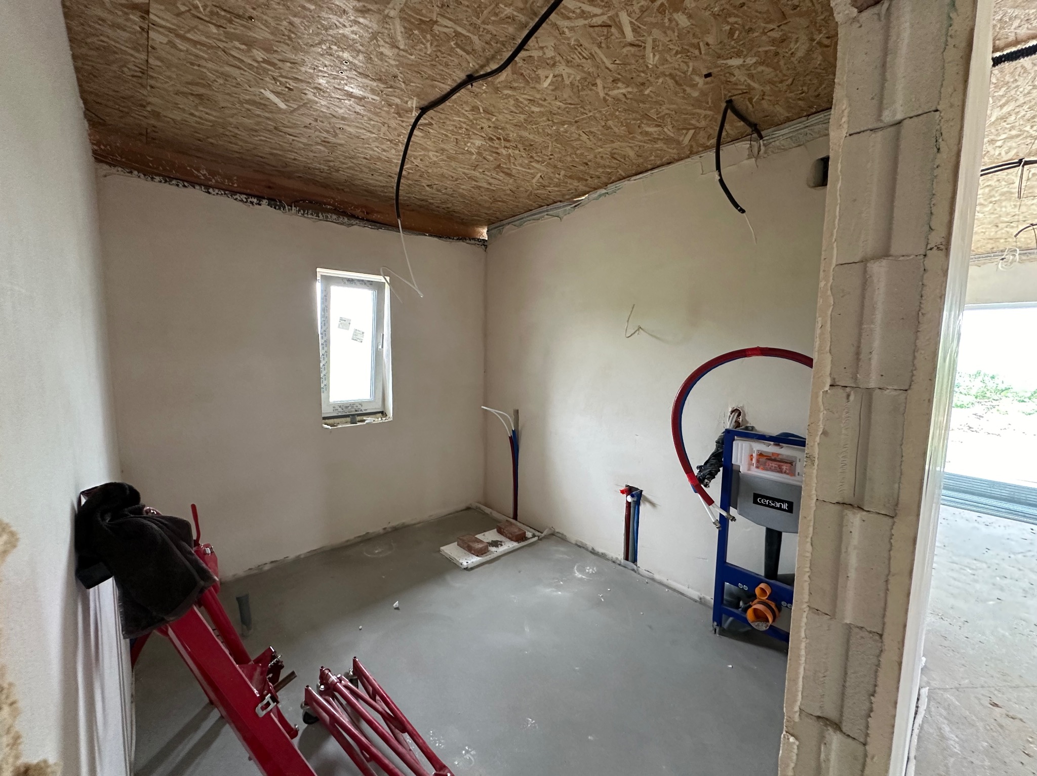 Wnętrze łazienki w trakcie prac wykończeniowych: ściany po nałożeniu gładzi, sufit z płyt OSB, widoczne instalacje wodne i stelaż podtynkowy WC marki Cersanit, na podłodze wylewka samopoziomująca...