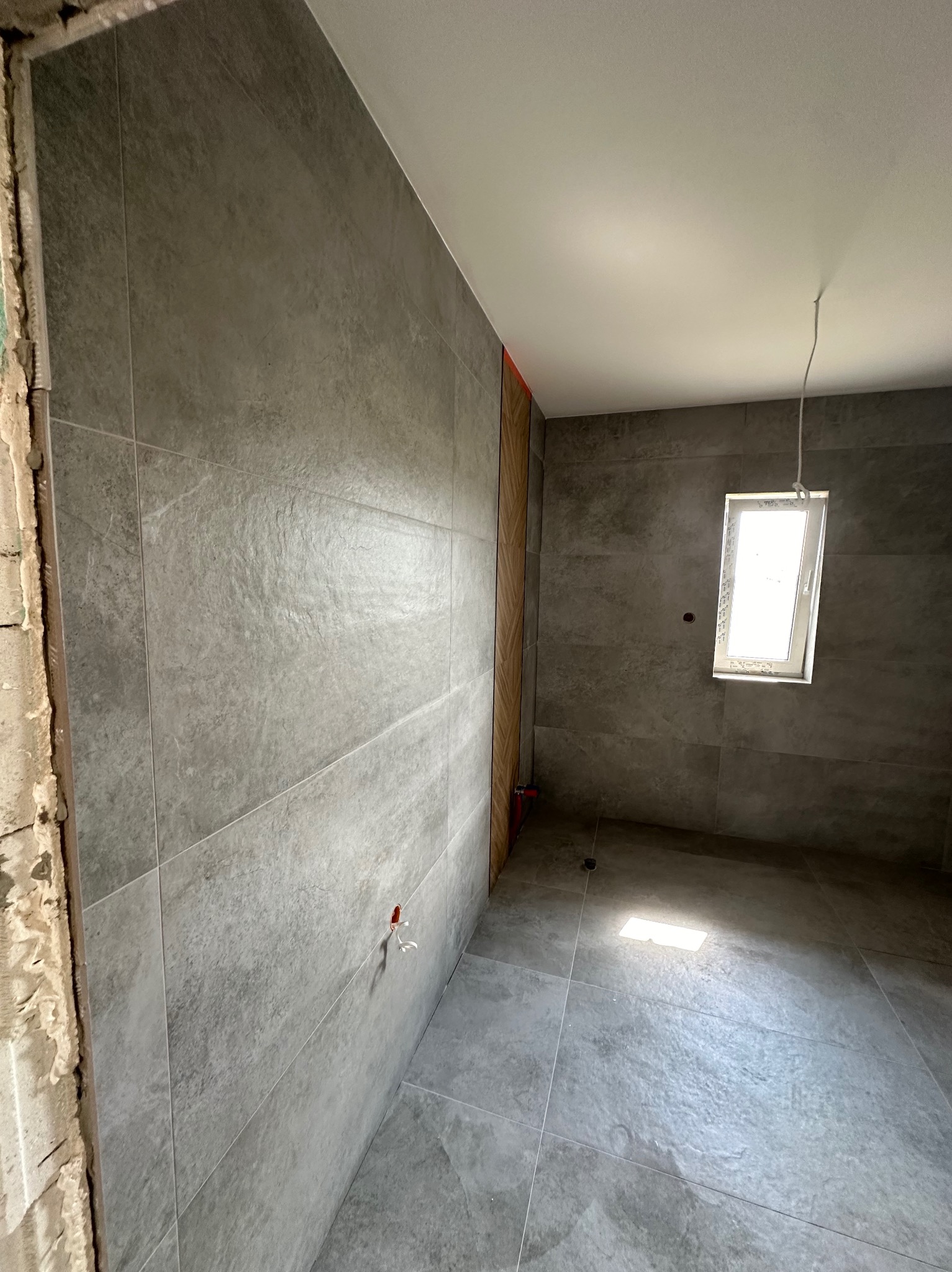 Szare płytki imitujące kamień na ścianach i podłodze łazienki w trakcie remontu, widoczne niedokończone obrzeża i wystające przewody.