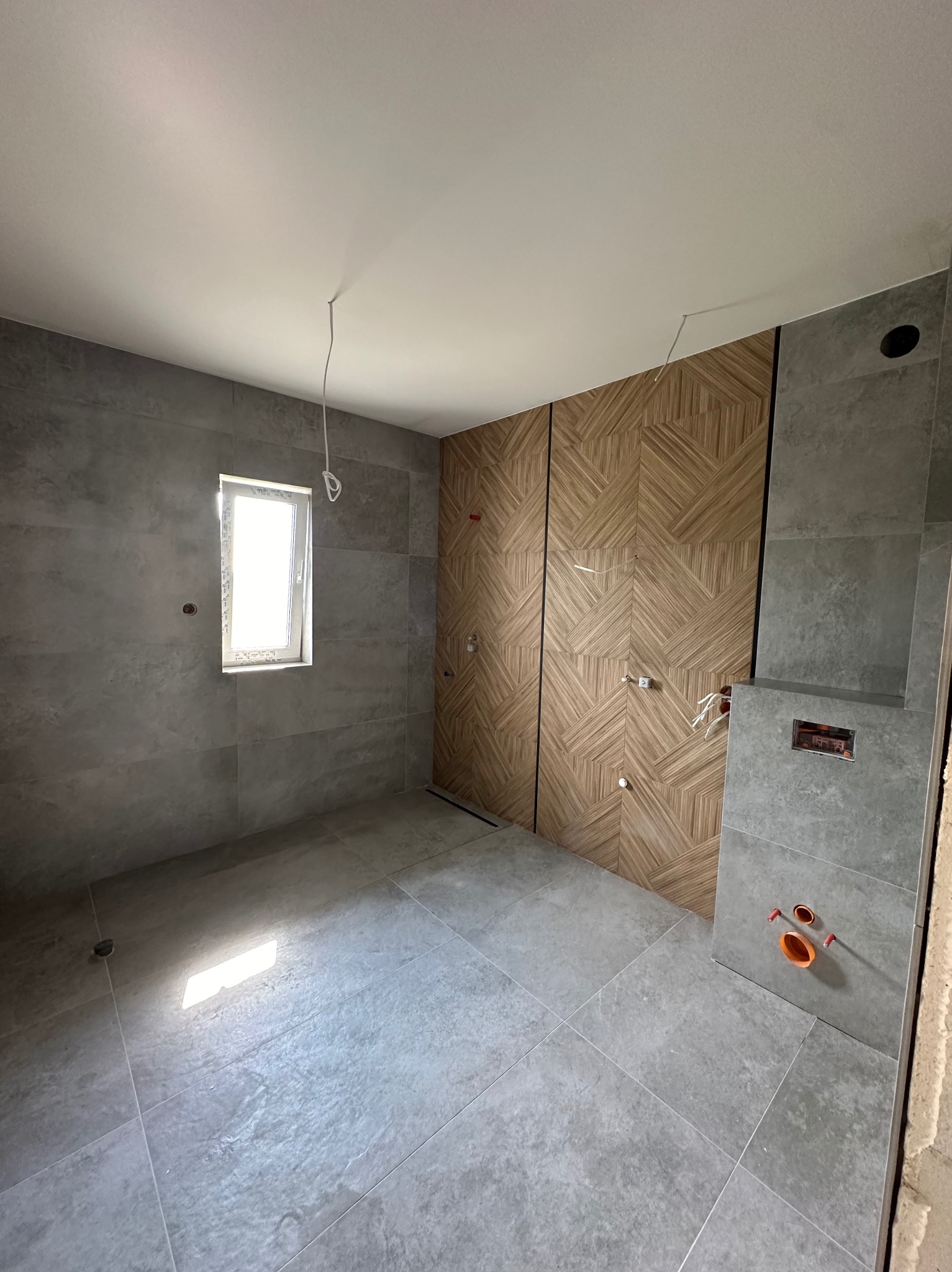 Wnętrze łazienki w trakcie wykańczania, widoczne szare płytki na podłodze i ścianach, dekoracyjna ściana z drewnopodobnych paneli z geometrycznym wzorem, instalacja podtynkowego WC z wystającymi...