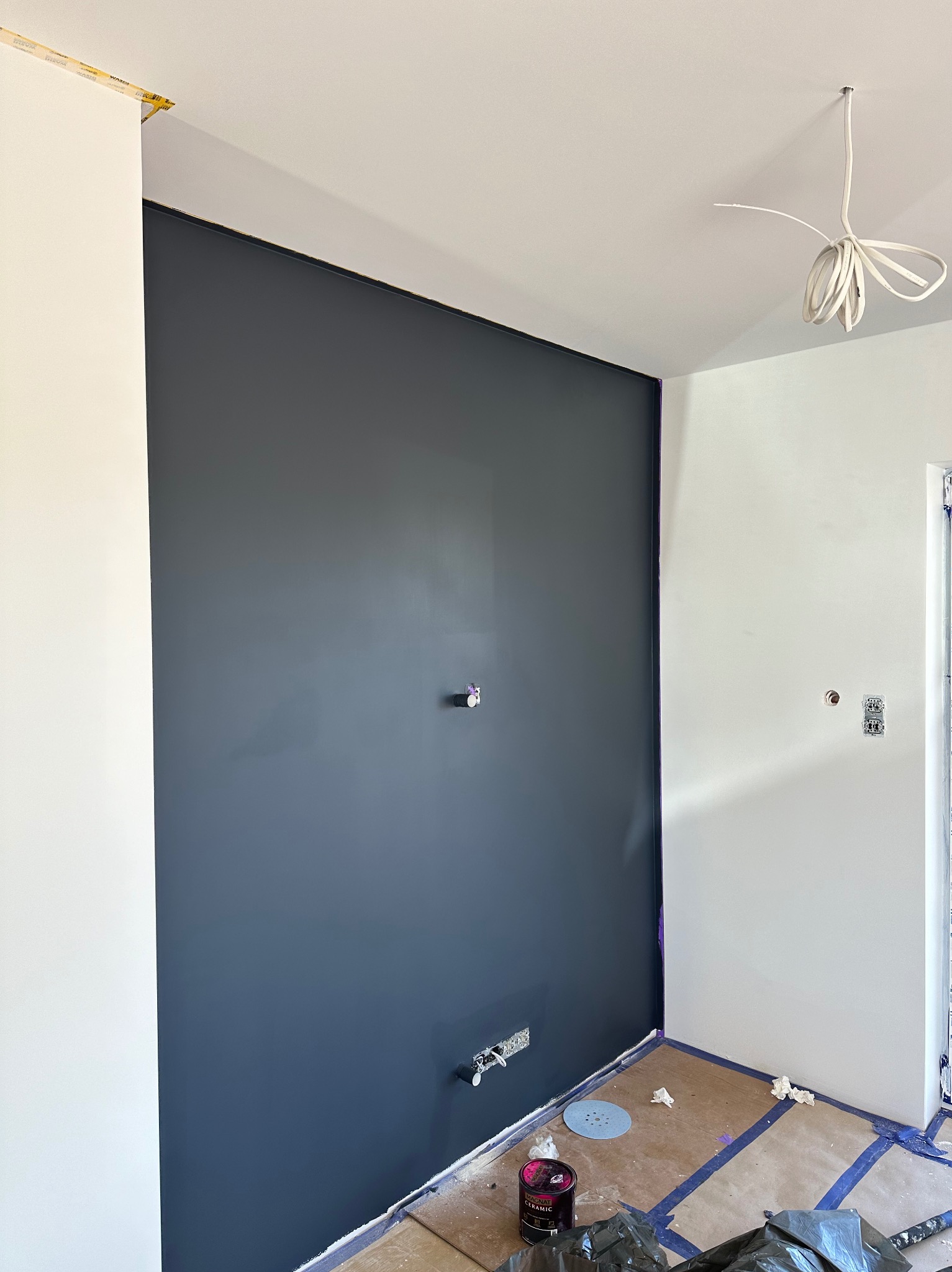 Świeżo pomalowana ściana na ciemny grafit w narożniku pokoju, z widocznymi puszkami elektrycznymi i zabezpieczoną podłogą, przygotowana do dalszych prac wykończeniowych.