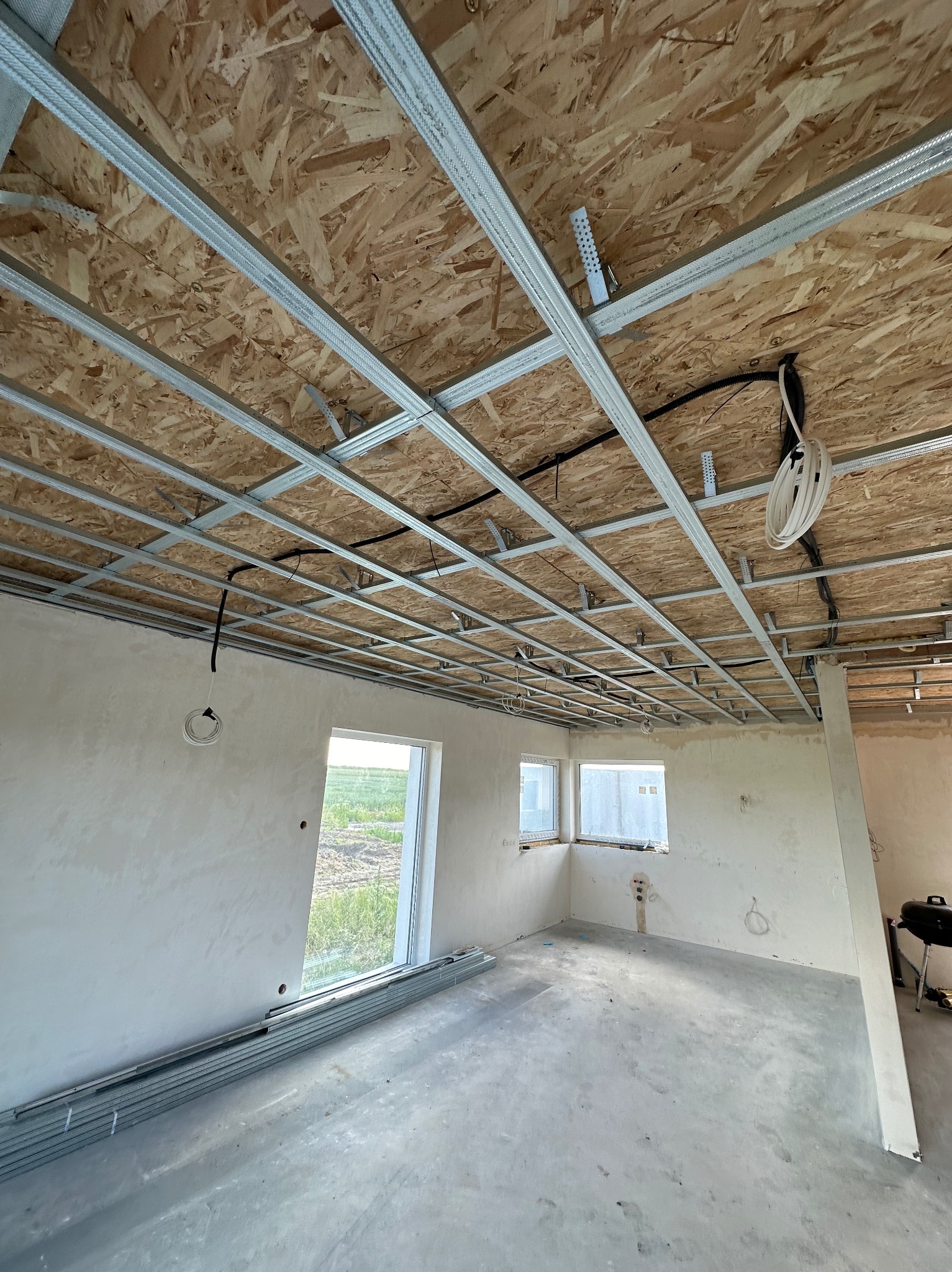Wnętrze w trakcie montażu metalowej konstrukcji sufitu podwieszanego na płycie OSB, widoczne kable elektryczne i przygotowane ściany do dalszych prac.