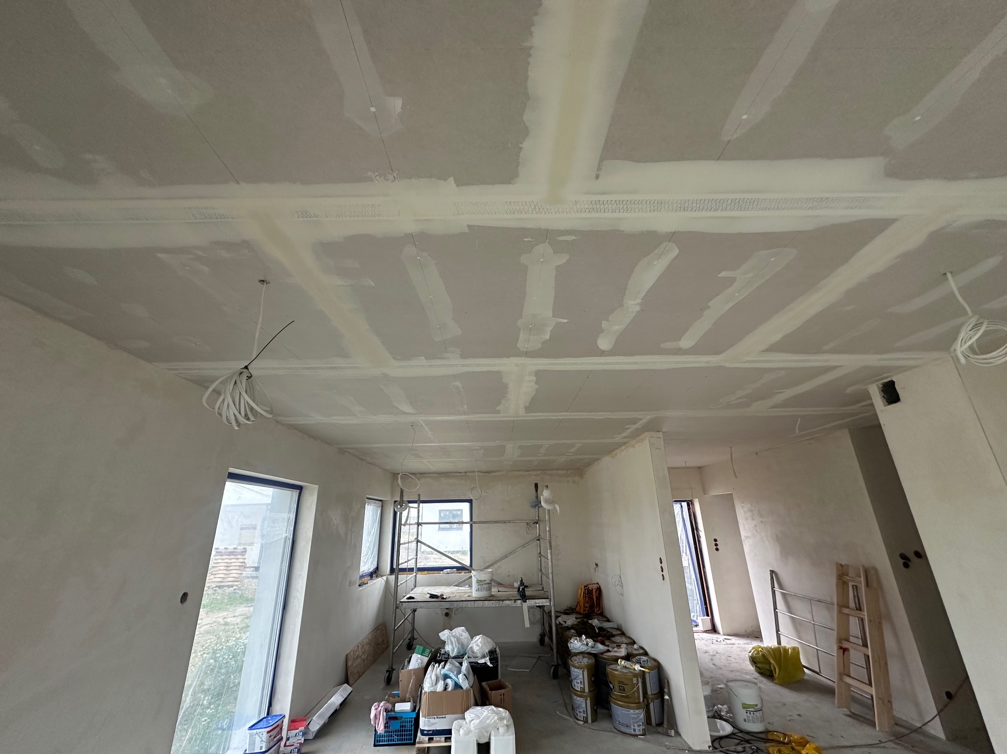 Wnętrze w trakcie prac wykończeniowych: sufit z płyt kartonowo-gipsowych ze świeżo nałożoną warstwą szpachli, widoczne łączenia wzmocnione siatką, rusztowanie z narzędziami i materiałami...