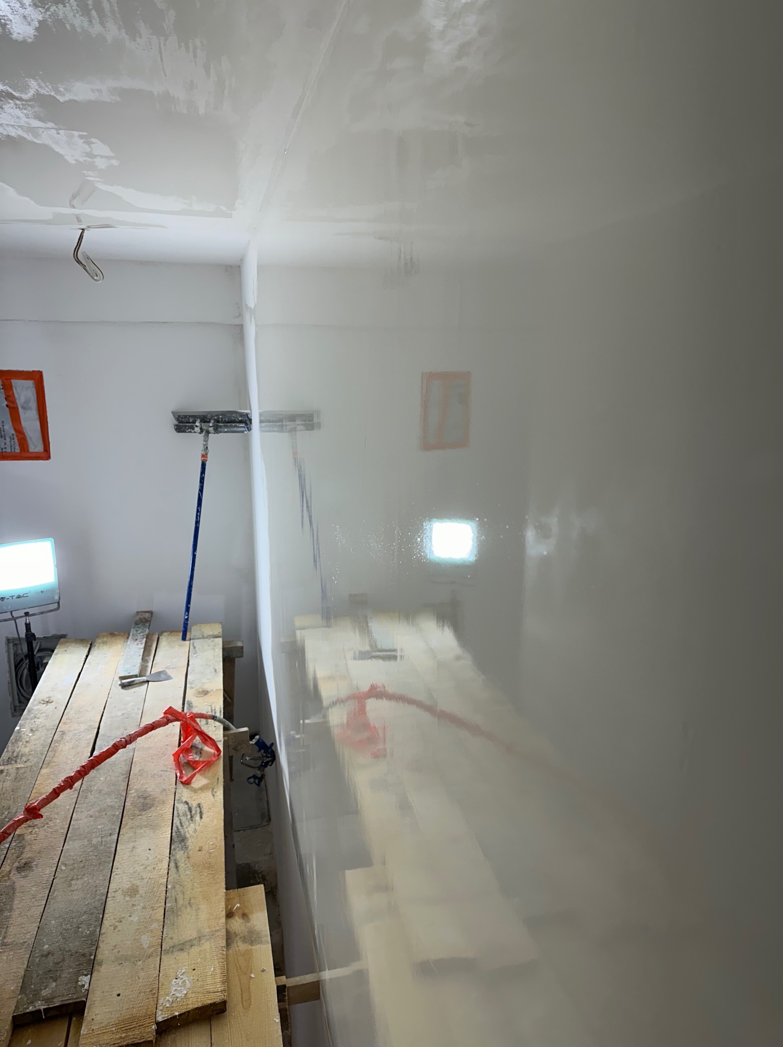 Wnętrze w trakcie prac wykończeniowych: świeżo nałożona, mokra warstwa gładzi na ścianach i suficie, widoczne rusztowanie robocze z narzędziami, lampa oświetleniowa i szpachla na kiju.