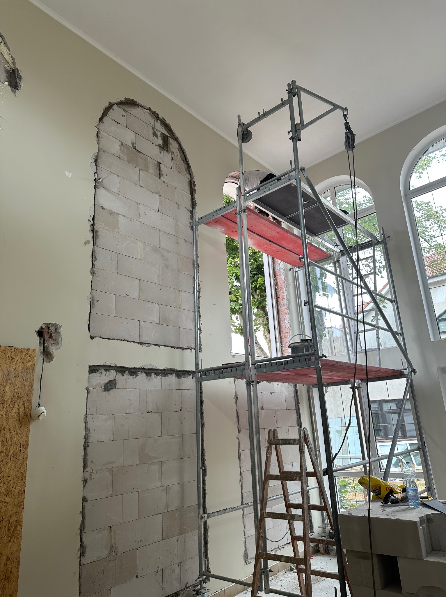 Wnętrze w trakcie prac murarskich: w ścianie wycięte otwory w kształcie łuków, widoczne bloki z betonu komórkowego, rusztowanie, drabina i narzędzia.