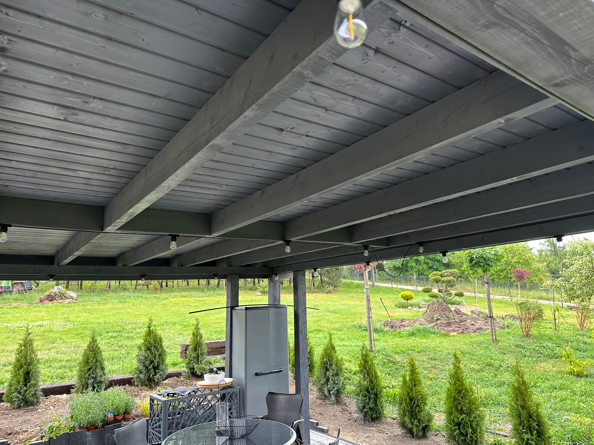 Szare drewniane zadaszenie tarasu z widokiem na ogród, oświetlone żarówkami, z elementami wyposażenia na pierwszym planie.