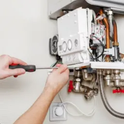 Serwisant z czerwoną rękojeścią śrubokręta reguluje biały, wiszący piec gazowy z miedzianymi rurkami i zaworami na tle ściany z delikatną fakturą.