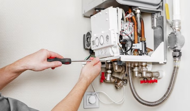 Serwisant z czerwoną rękojeścią śrubokręta reguluje biały, wiszący piec gazowy z miedzianymi rurkami i zaworami na tle ściany z delikatną fakturą.